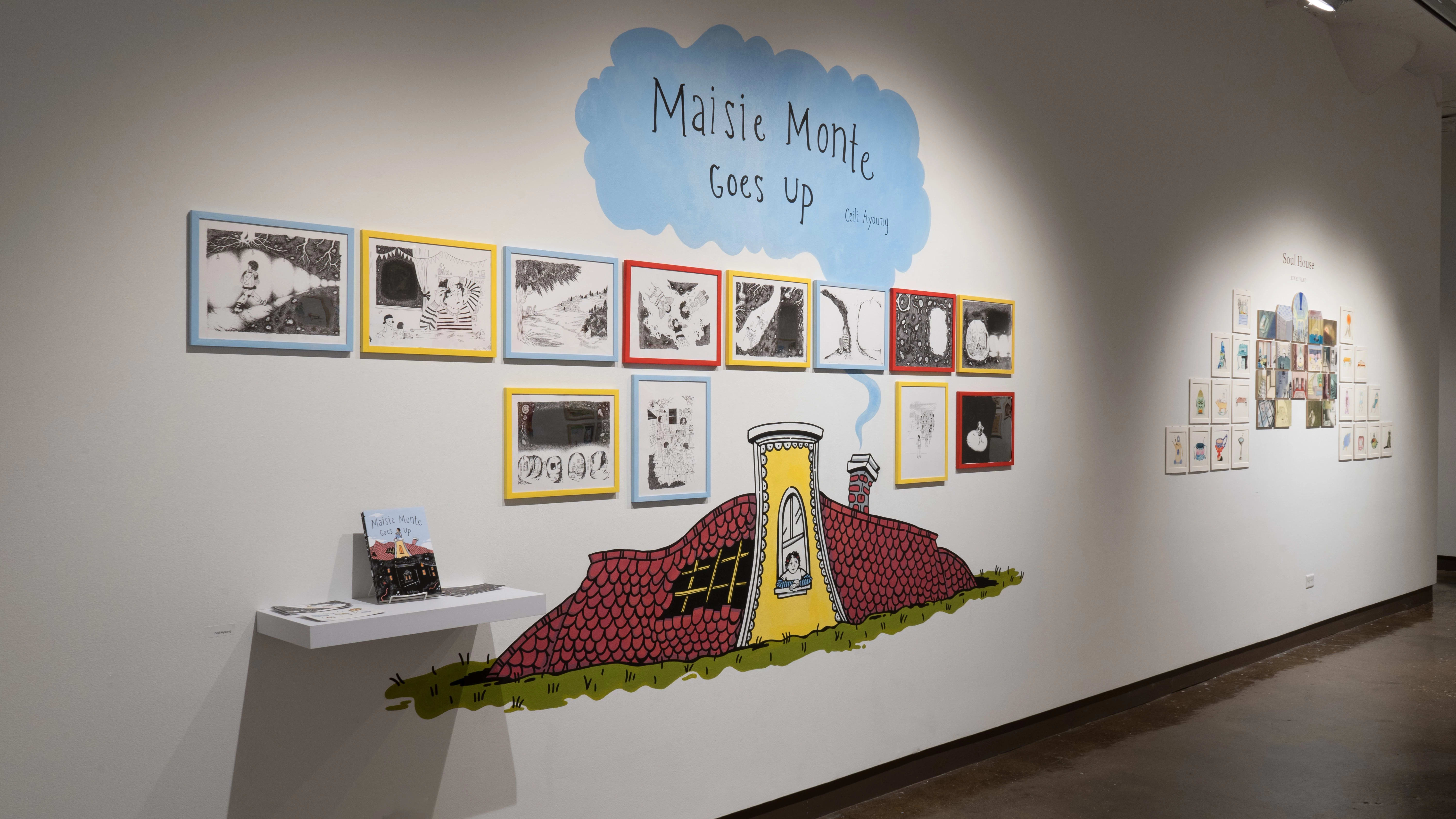 Black and white illustrations are framed in blue, yellow, and red frames and hung in two rows on a wall of gallery three. On the wall above the frames, “Maisie Monte Goes Up” is painted on a blue cloud, and below them, a small cottage is painted.