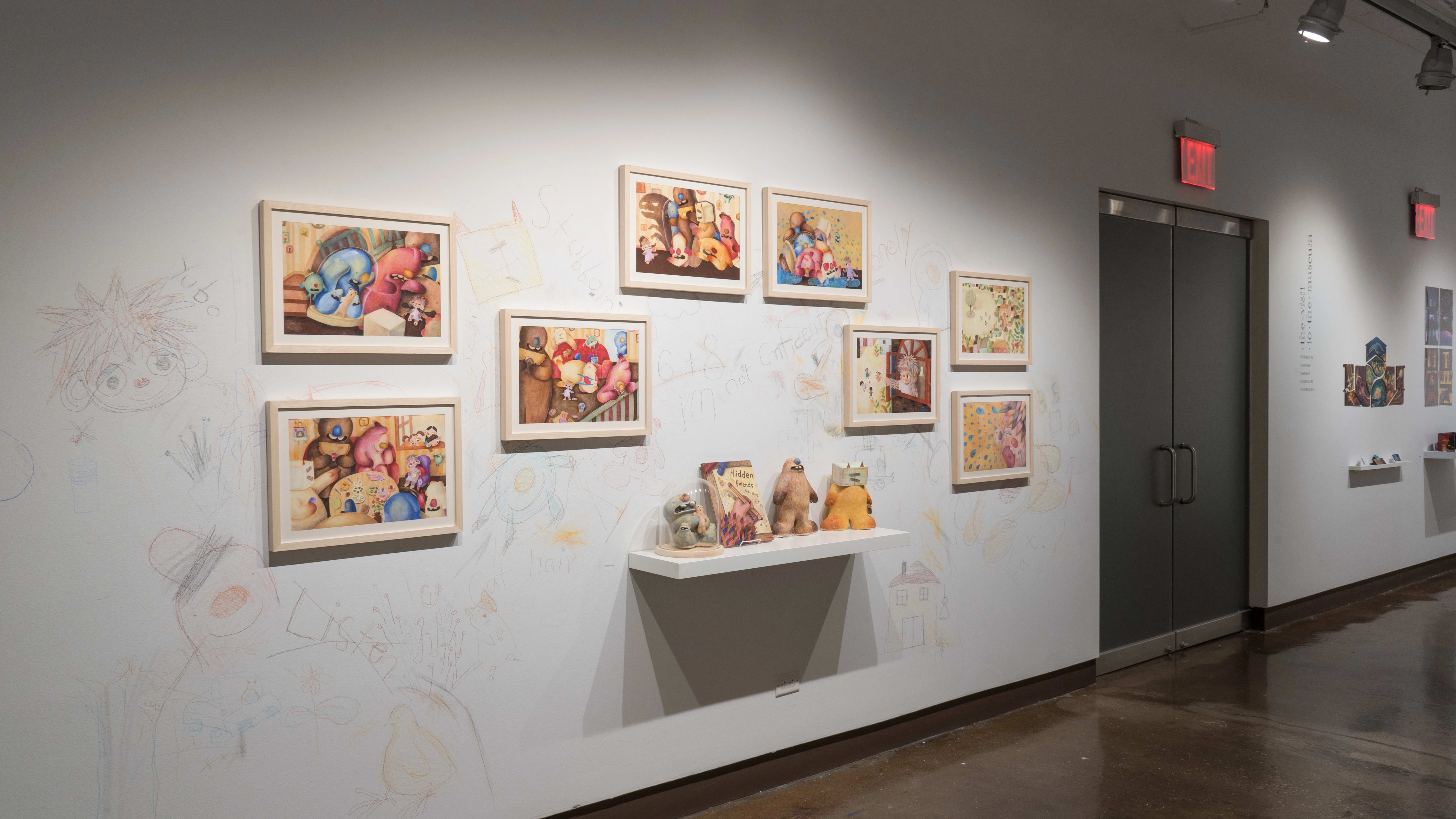 In the left hallway leading from gallery three to gallery four, colorful, framed illustrations are hung on the left wall. A shelf below them holds sculptures of the illustrated characters and a book. The surrounding wall is sketched on with colored pencils.