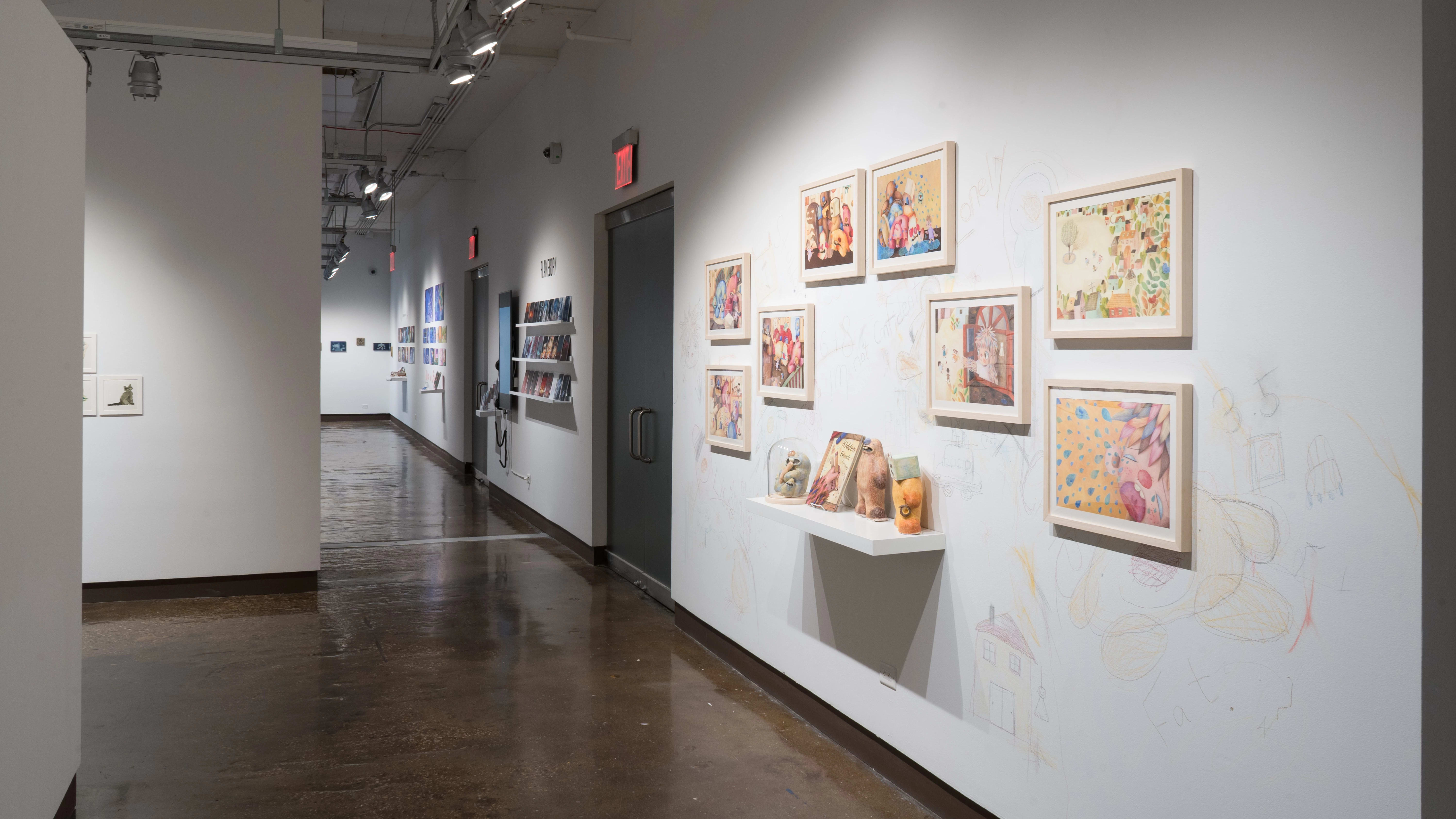 In the left hallway leading from gallery three to gallery four, colorful, framed illustrations are hung on the left wall. A shelf below them holds sculptures of the illustrated characters and a book. The surrounding wall is sketched on with colored pencils.