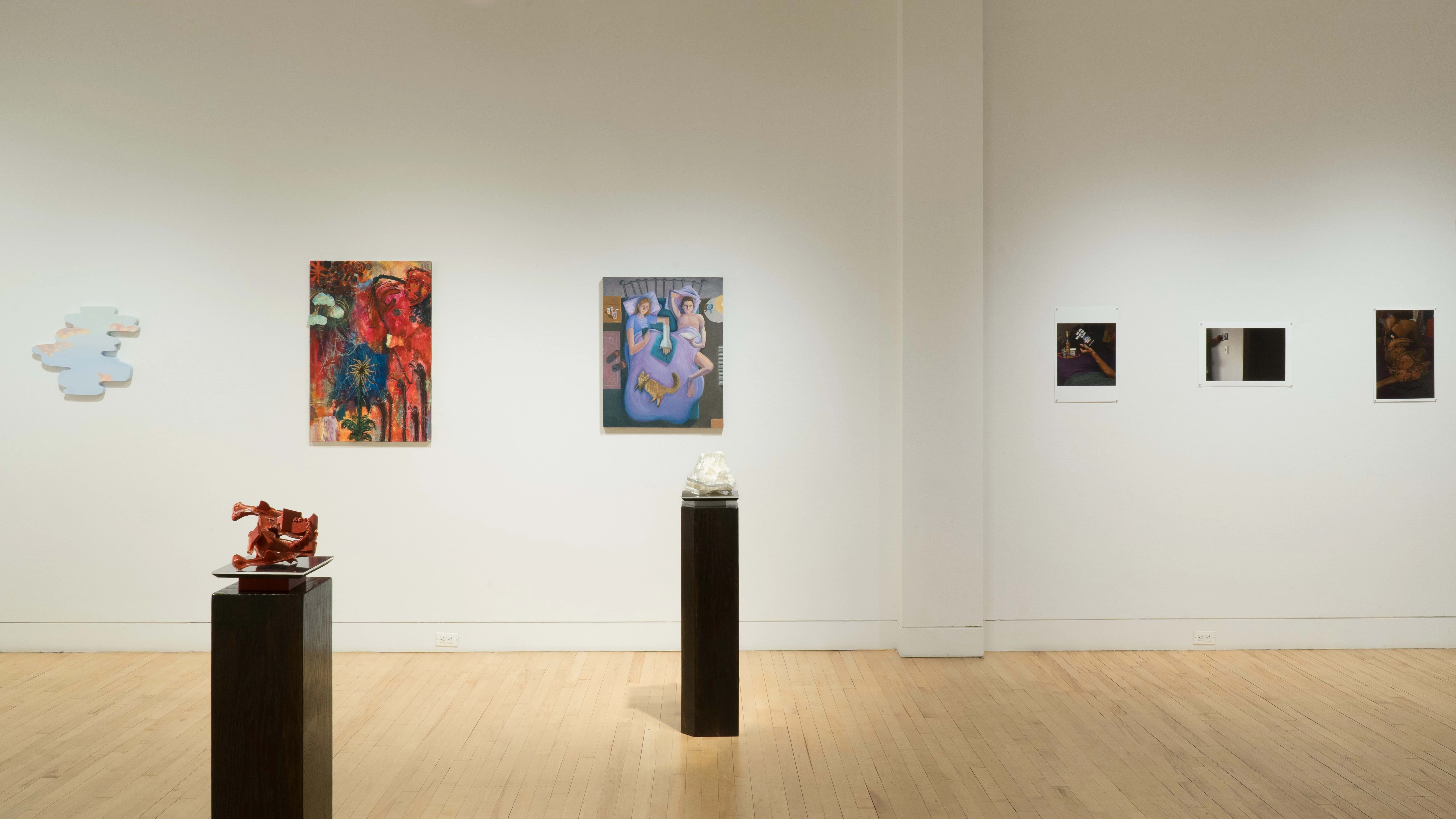 In the center of the gallery, a red sculpture and a white sculpture are placed on their own black pedestals. On the left half of the wall beyond them, three colorful but varied paintings hang beside each other. On the right half, there are three shadowy photographs pinned to the wall.