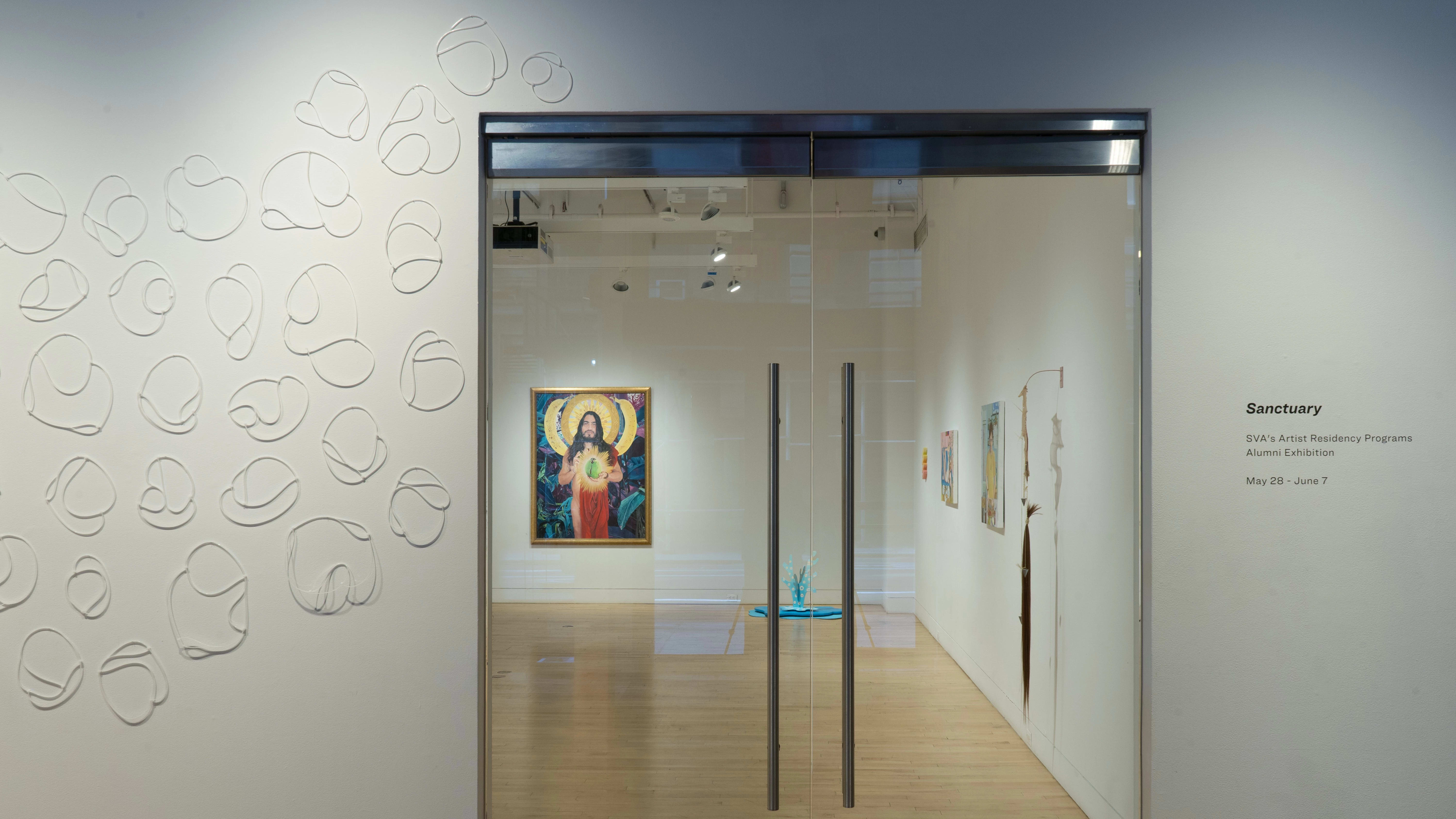 Facing the glass doors looking into the gallery, a painting of a saint or other holy figure hangs on the back wall. Sculptures and other paintings are also visible. Outside the door, zip ties are placed in a design on the wall.