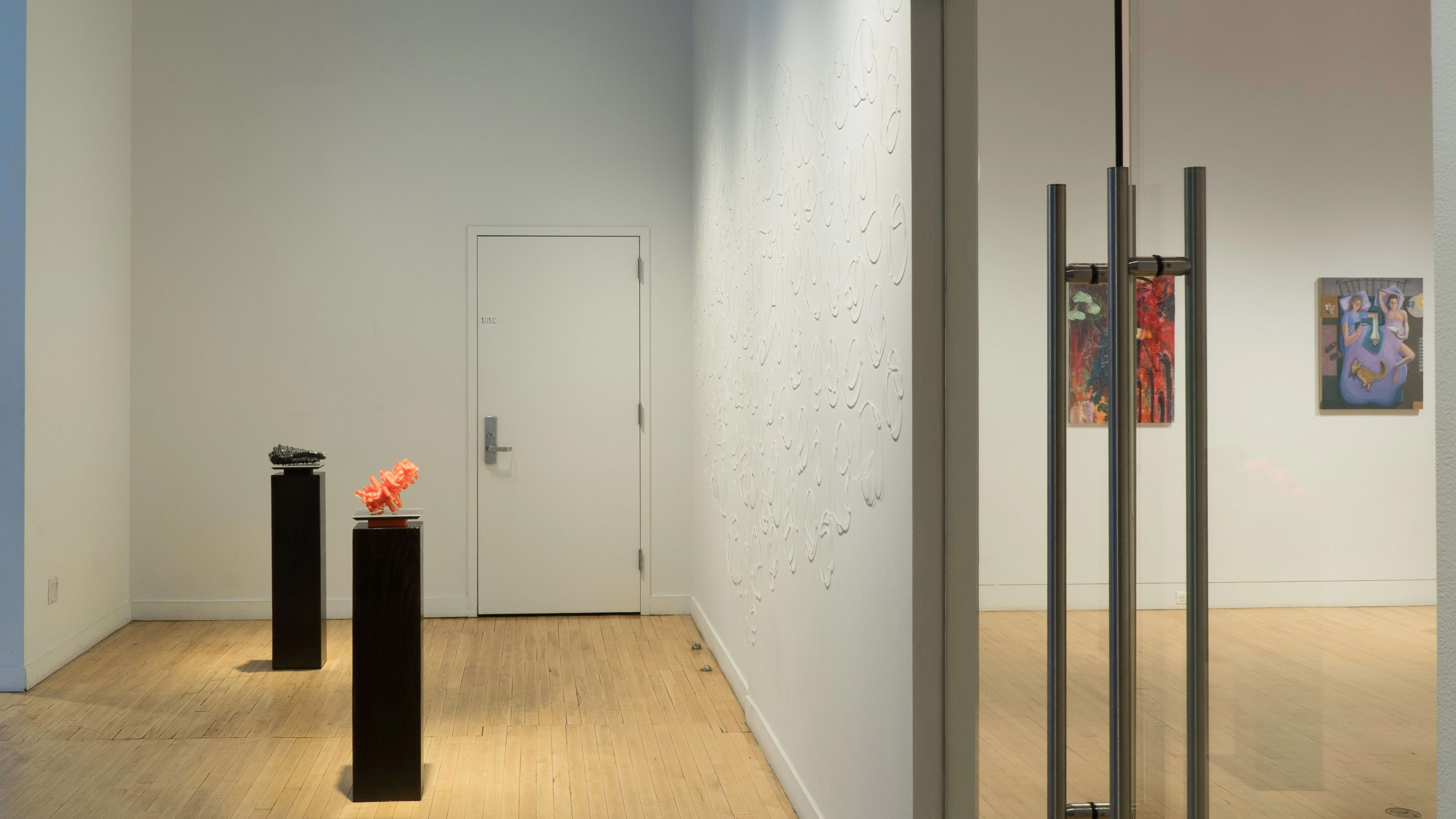 In the alcove of the SVA Flatiron Gallery, zip ties formed into loops are placed in a design all over the wall. In the center, an orange and a black sculpture sit on two black pedestals. Through the glass doors to the gallery, a few paintings are visible.