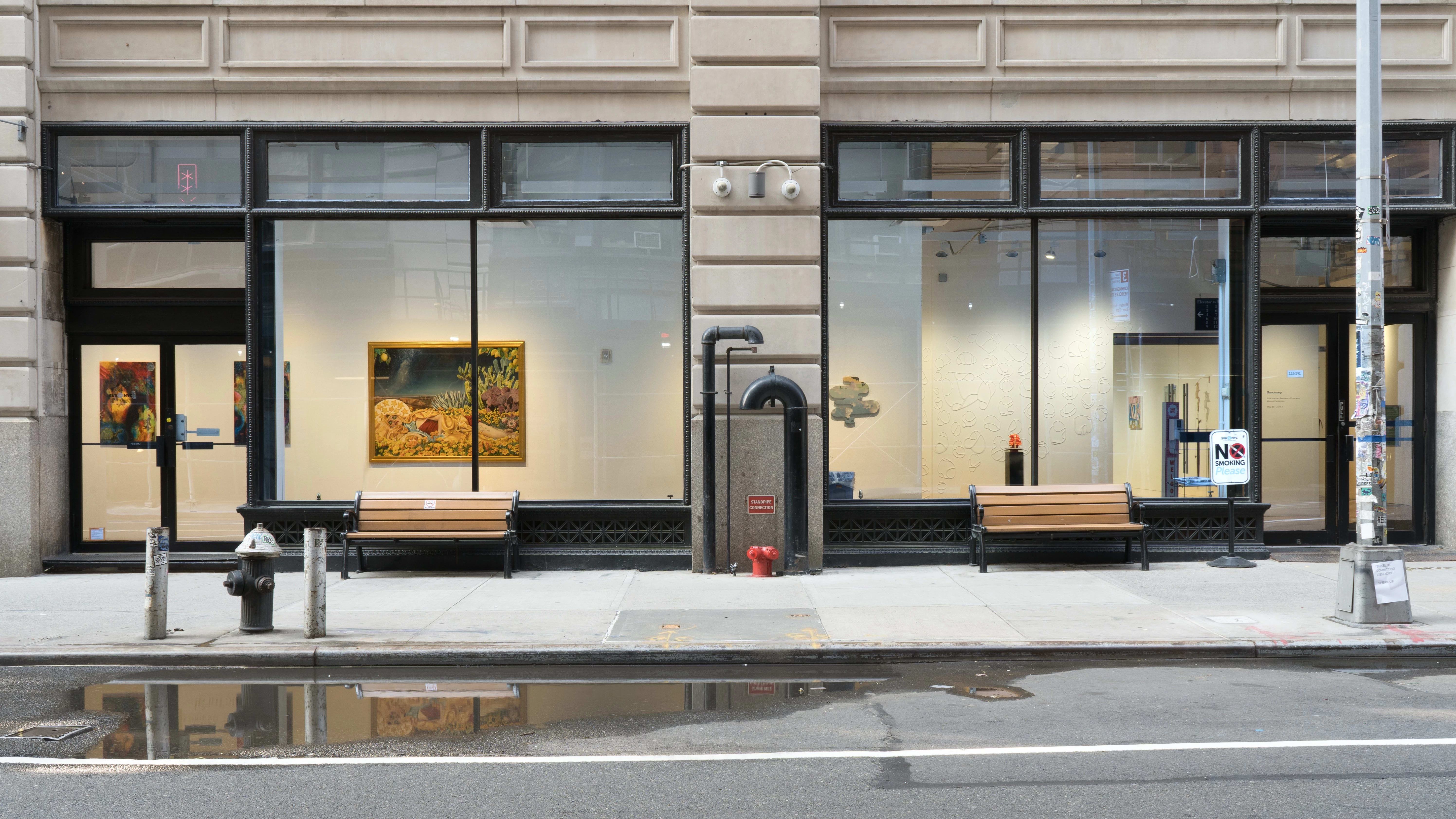 From the street outside a building, the SVA Flatiron Gallery and works hung in the hallway are visible through large windows. A few medium-large rectangular paintings hang beside a panel cut into an organic shape and painted with the colors of the sky.
