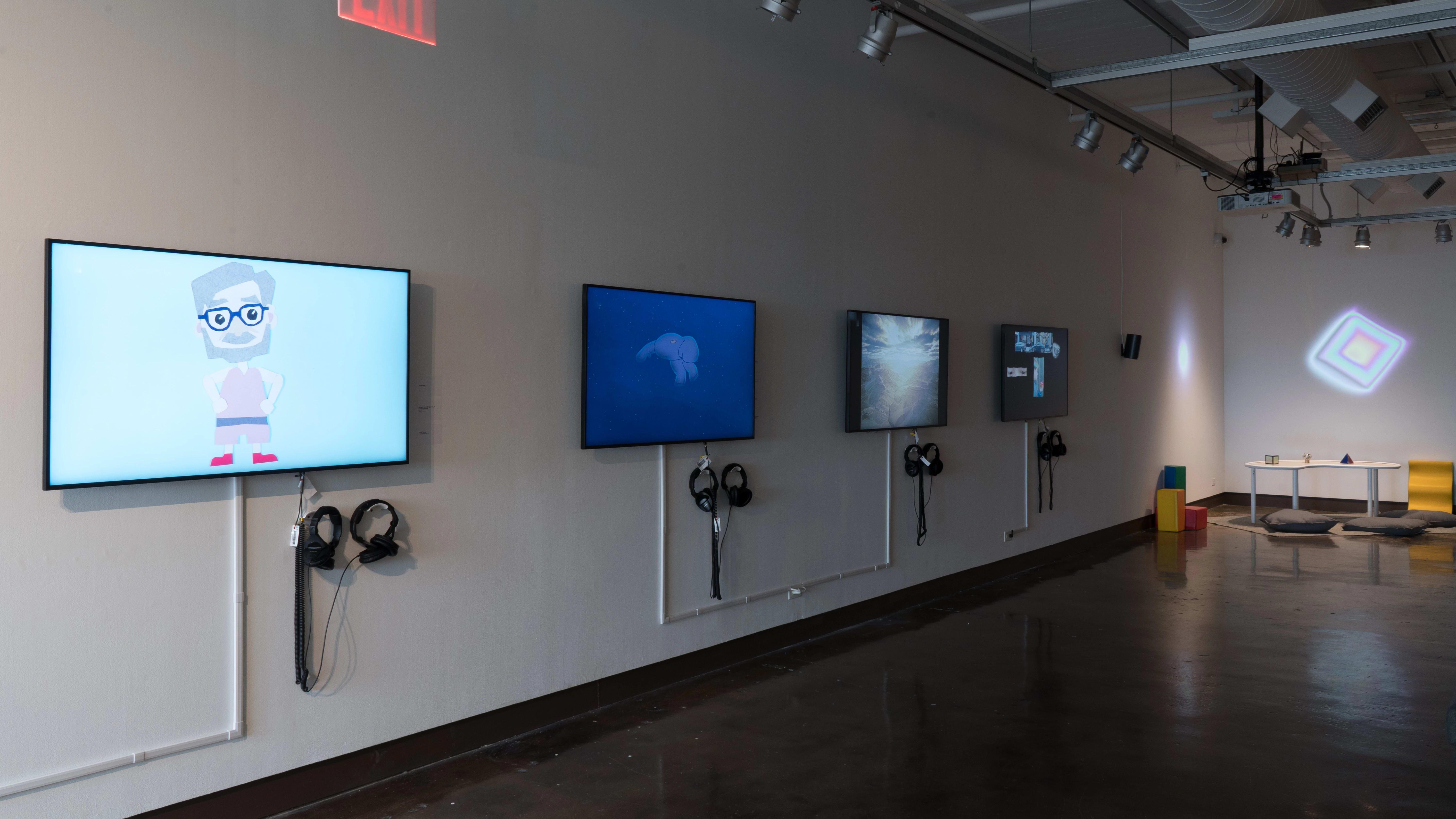 Four monitors along a wall in gallery one play many animated videos on a loop. In the corner to the right, there are colorful projections on the wall, and a small table with three differently shaped objects.