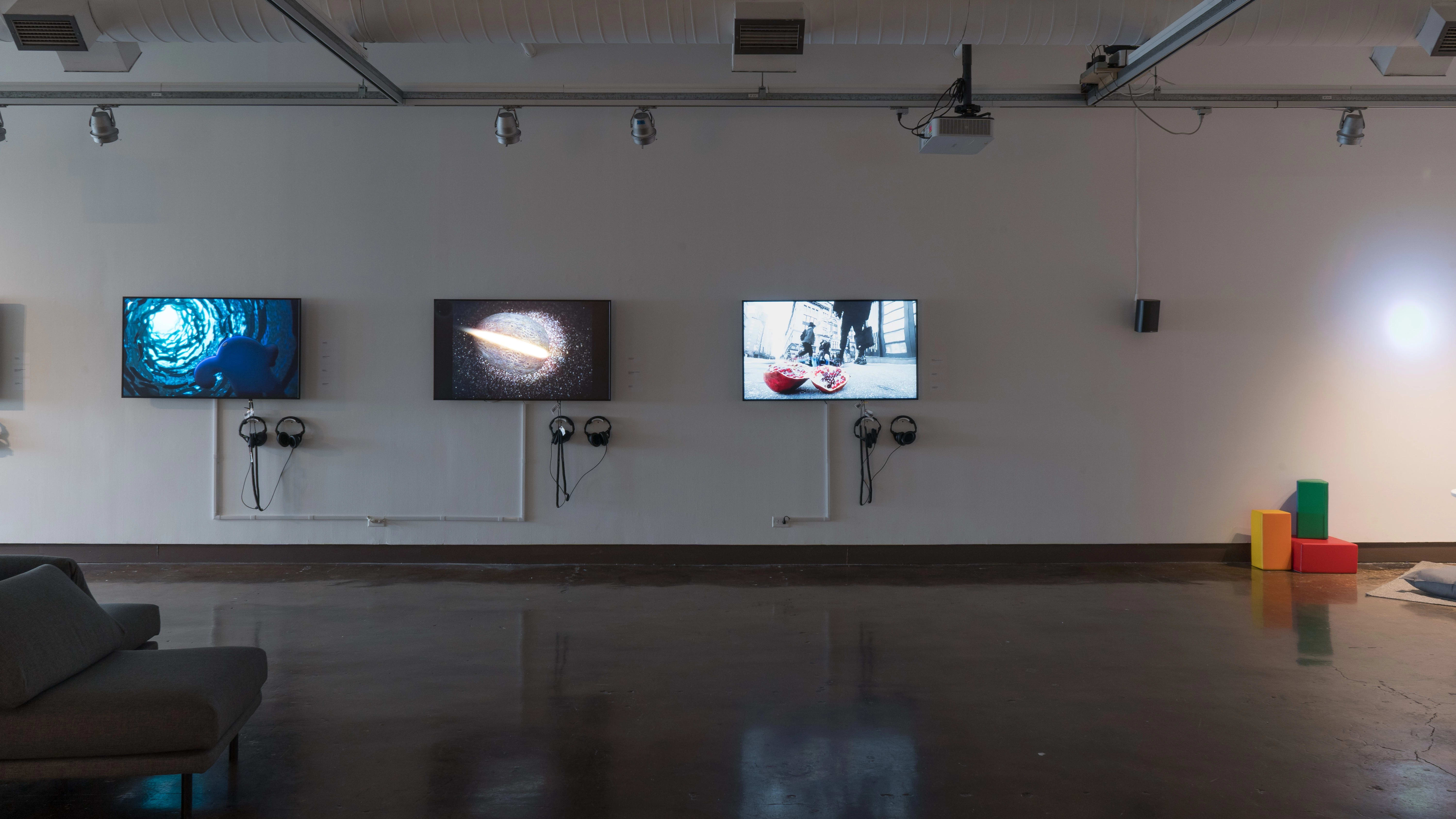 Three monitors along a wall in gallery one play many animated videos on a loop. Colorful blocks sit on the floor to the far right.