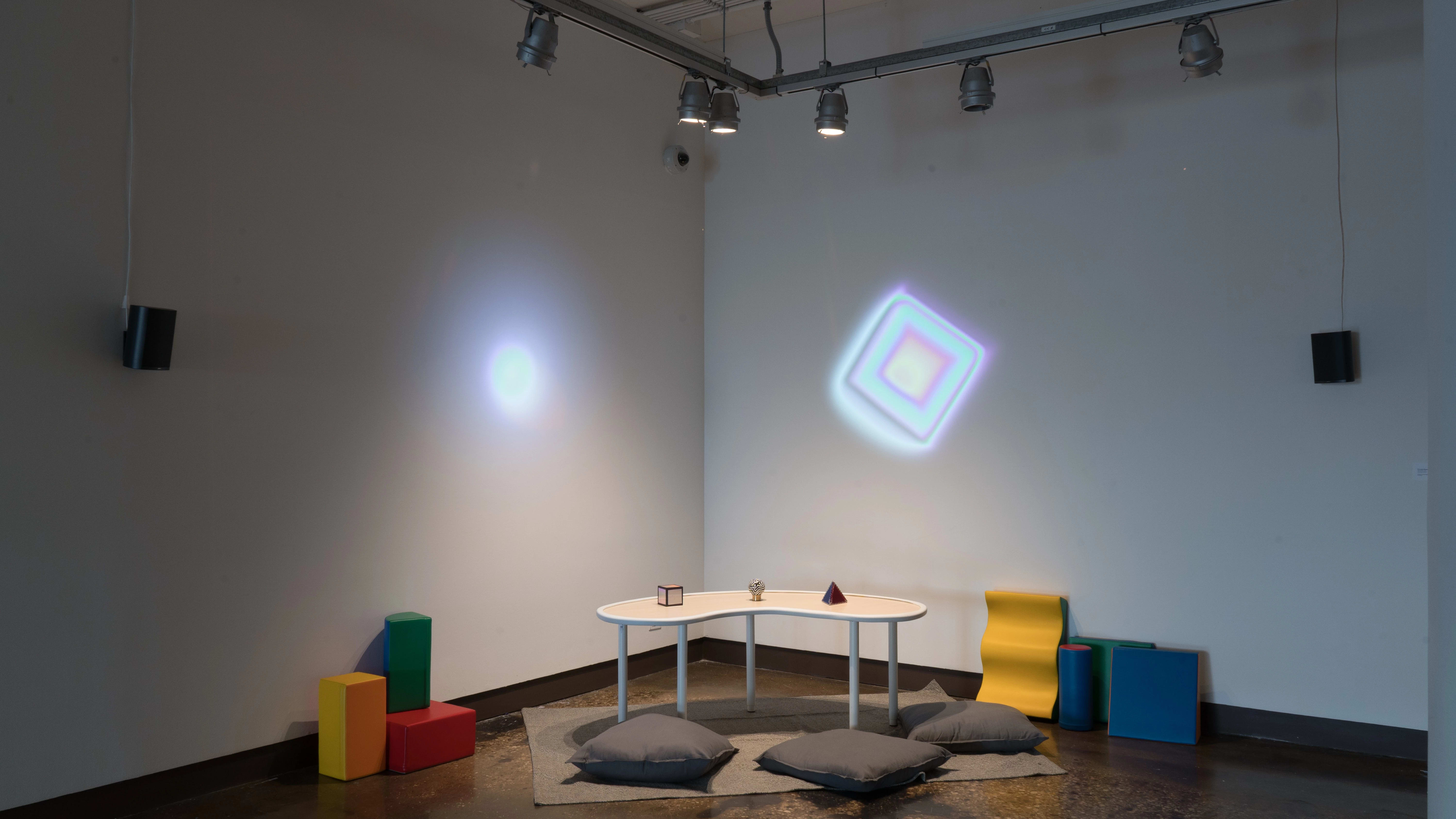 In a corner of gallery one, three objects (a cube, a sphere, and a pyramid) are laid out on a small table. The table is surrounded by floor pillows, and colorful projections glow on both walls.