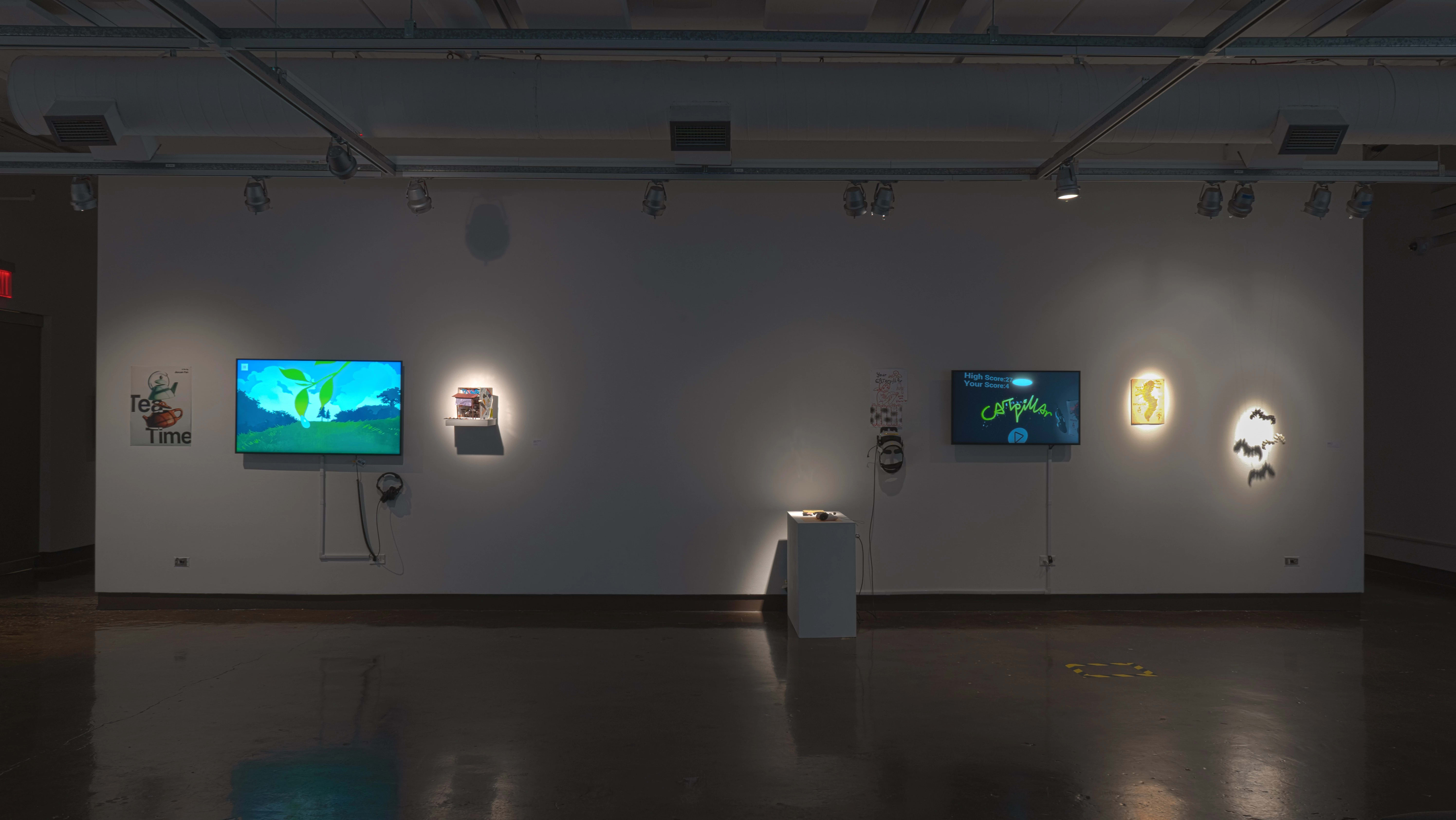 In gallery one, a monitor playing an animated video is flanked by a small poster and a shelf holding a small diorama. To the right, a VR video game is set up on a monitor, with a print and sculpture of caterpillars beside it.