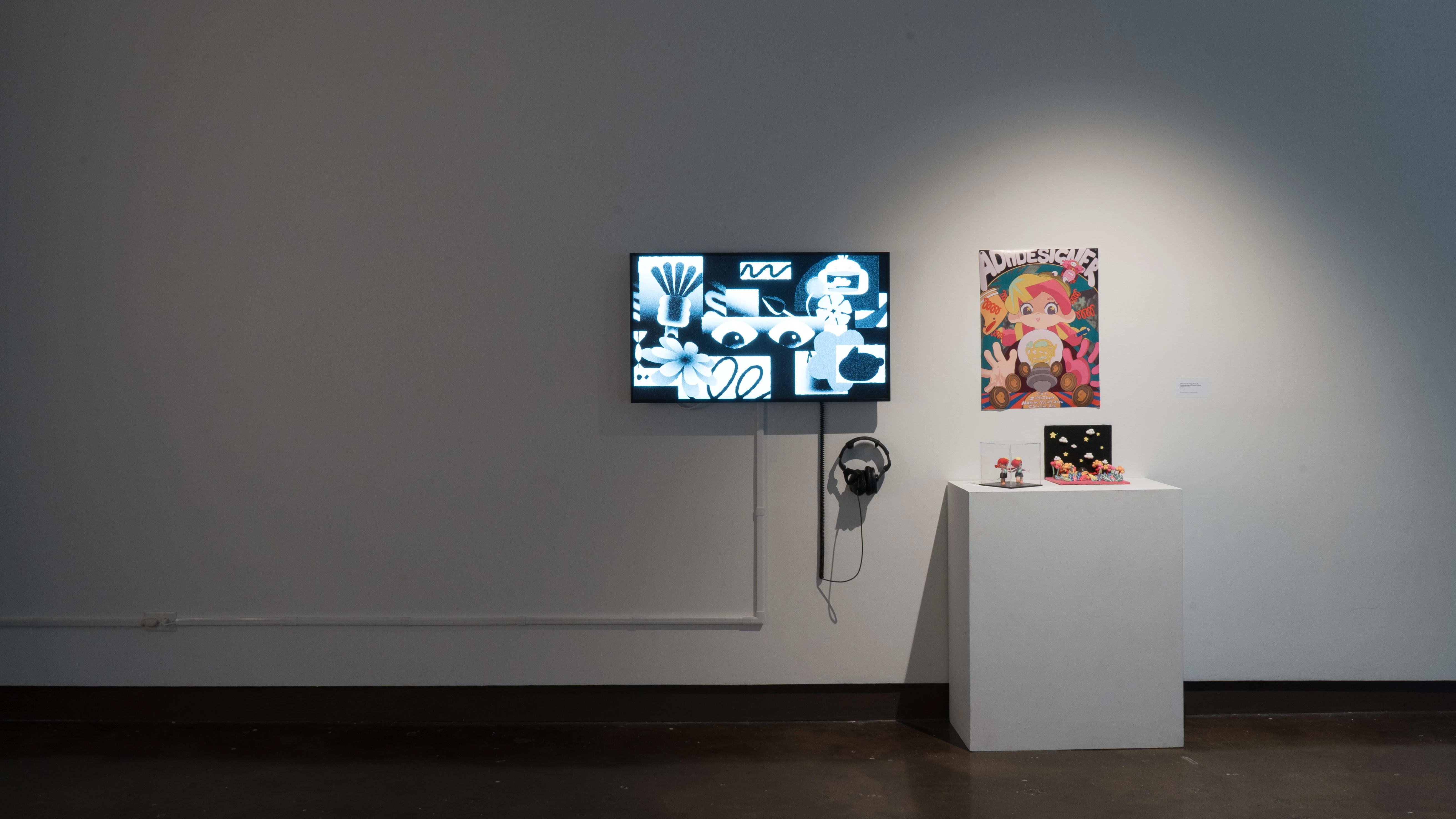 Beside a monitor playing an animated video, a white pedestal holds a small diorama and a tiny doll. A poster behind it depicts a girl with pink hair.