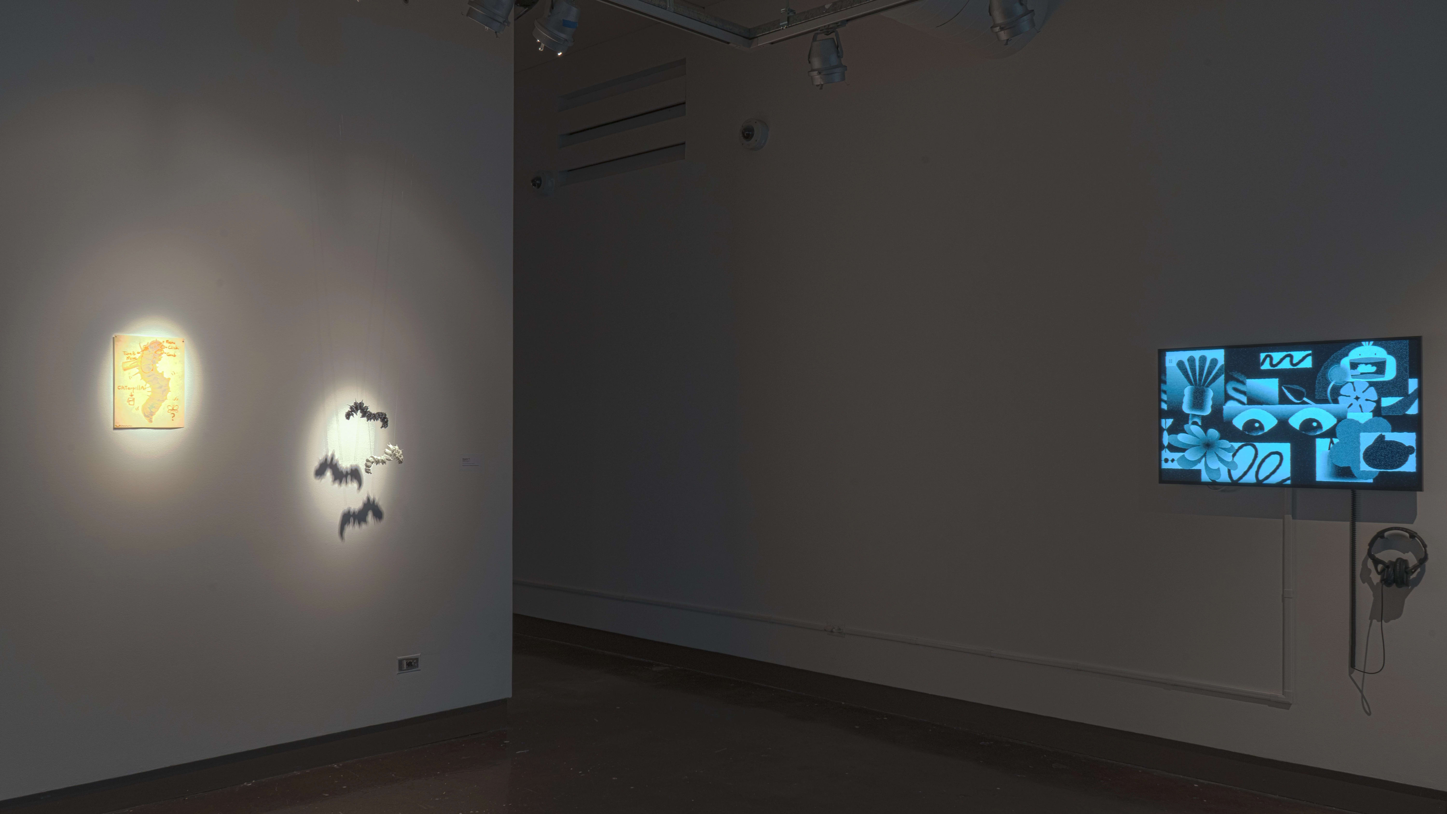 In a corner of gallery one, spotlights are focused on a print and a hanging sculpture of a caterpillar. To the right, a monitor plays an animated video.