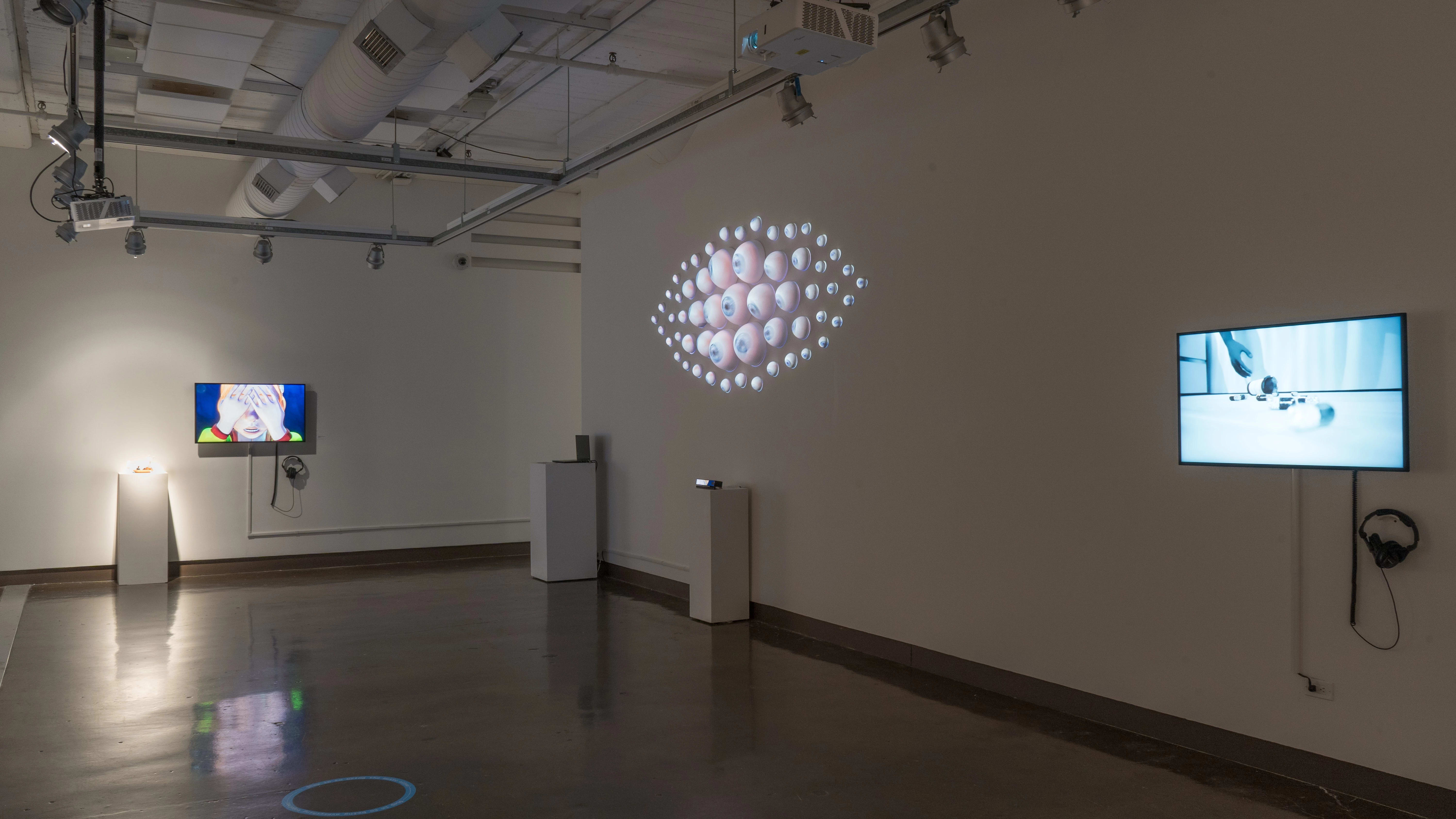 Many eyeballs of various sizes are arranged in an eye-like shape, protruding from the wall in gallery two. To the left and right of the eye, there are TV monitors playing animated videos.