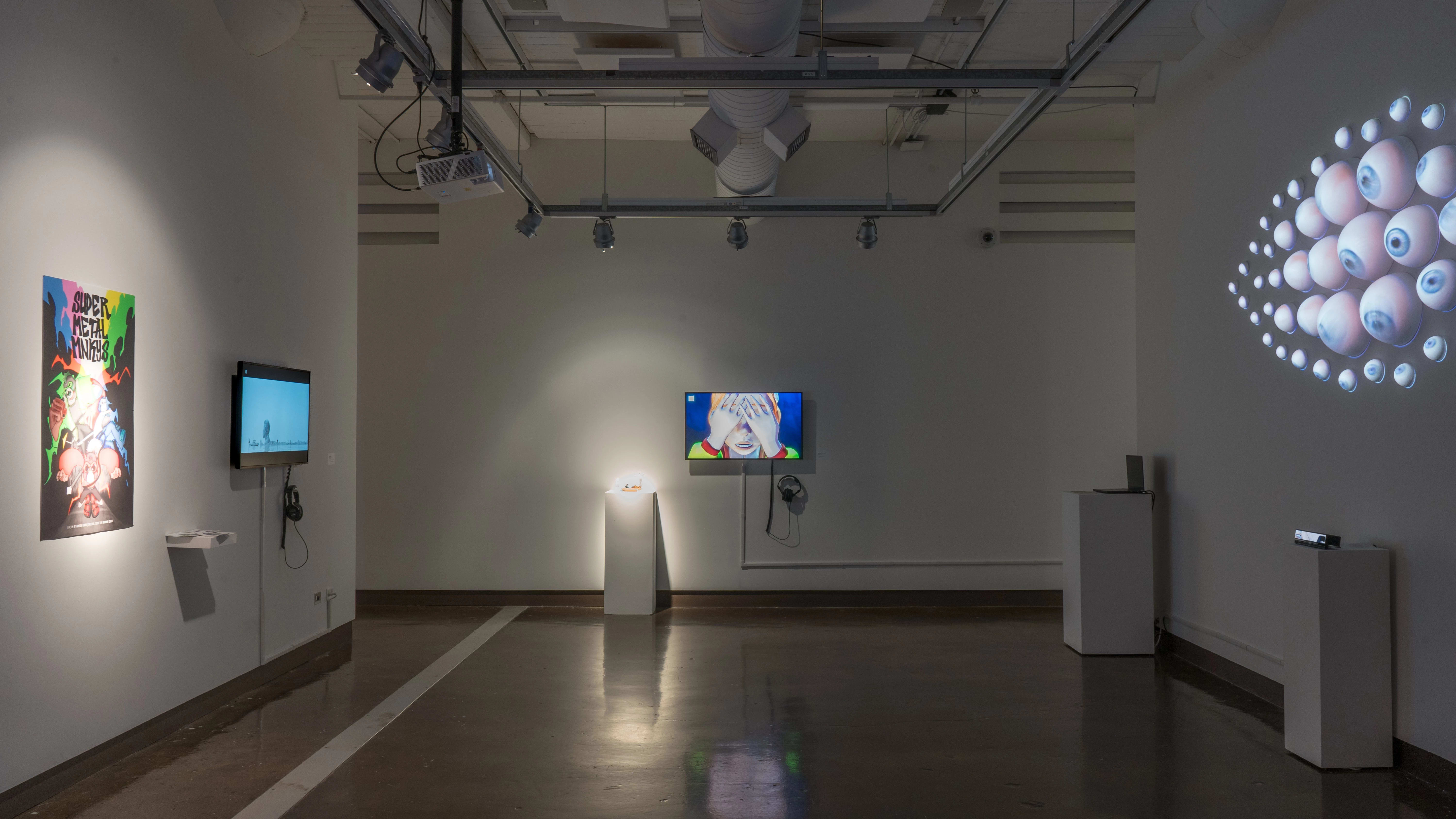 In gallery two, TV monitors hang on two of the three visible walls. On the third wall, many eyeballs of various sizes are arranged in an eye-like shape, protruding from the wall.