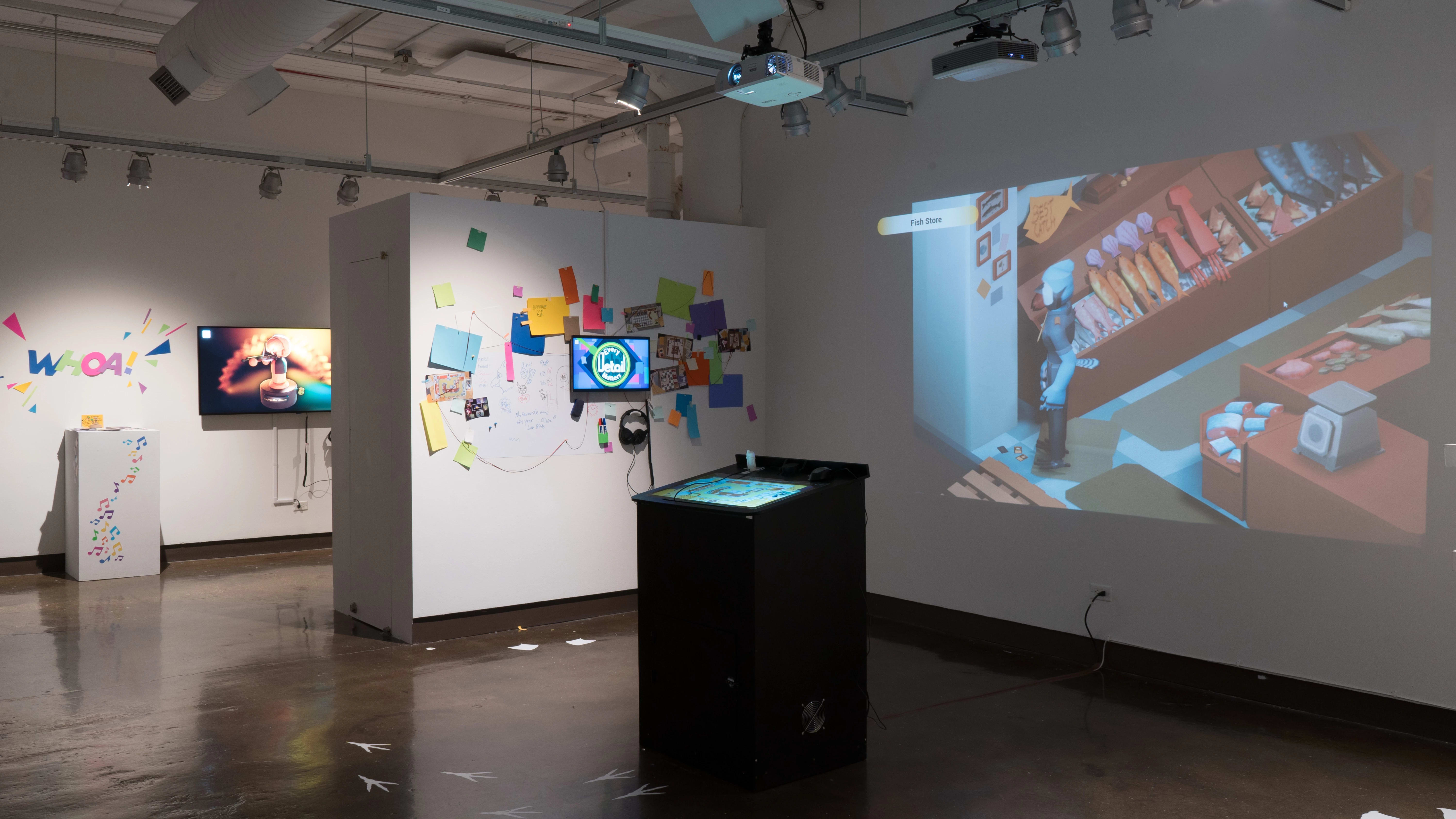 A small kiosk sits in the center of the gallery three alcove as a video game is projected on the wall behind it. On the side wall, a TV monitor plays an animated video, and many pieces of colorful paper surround the monitor.