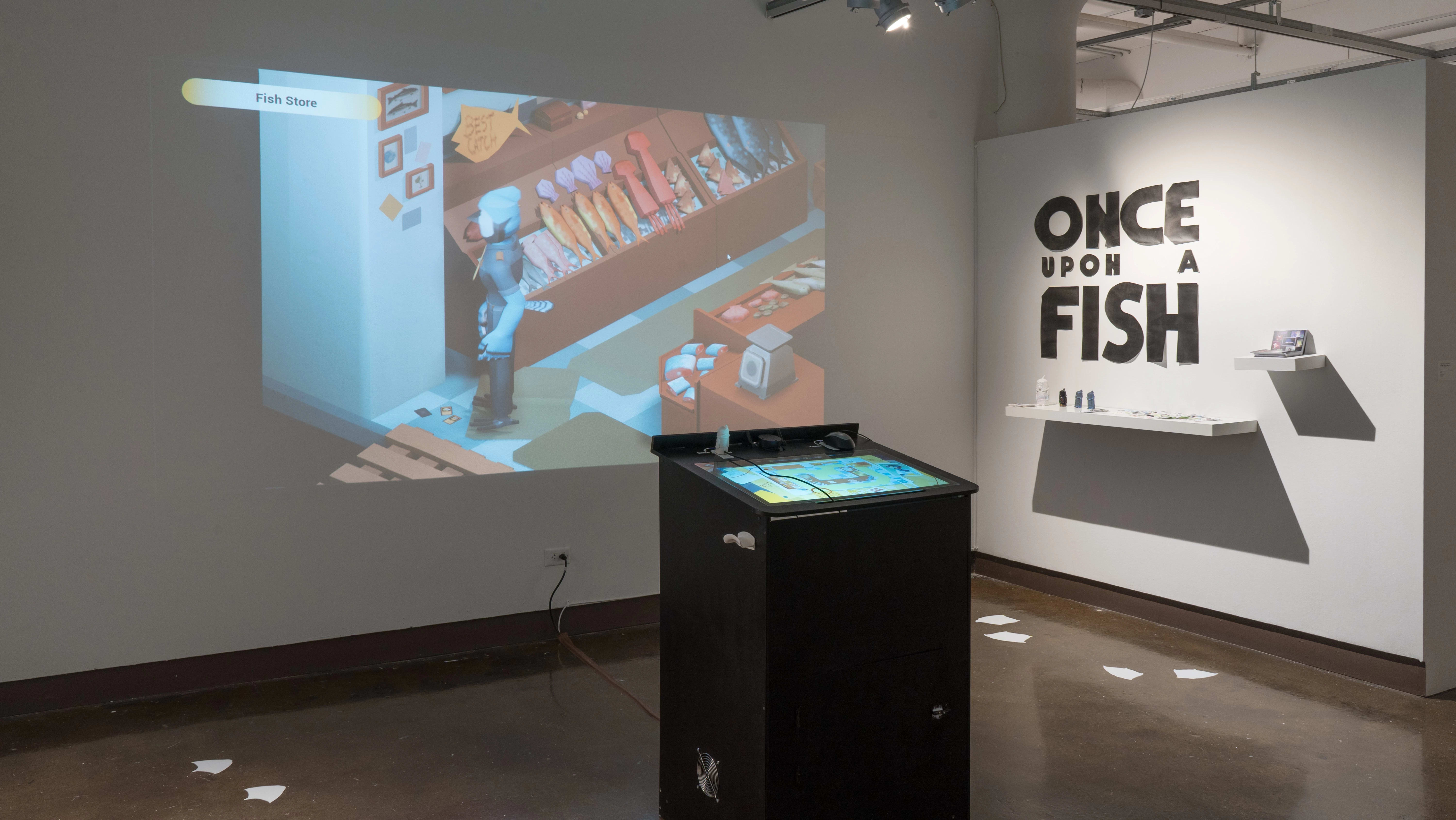 In the gallery three alcove, bold text on one wall reads “Once Upon A Fish.” A black kiosk in the center of the space has a screen to play a video game. The game is projected onto the wall behind the kiosk.
