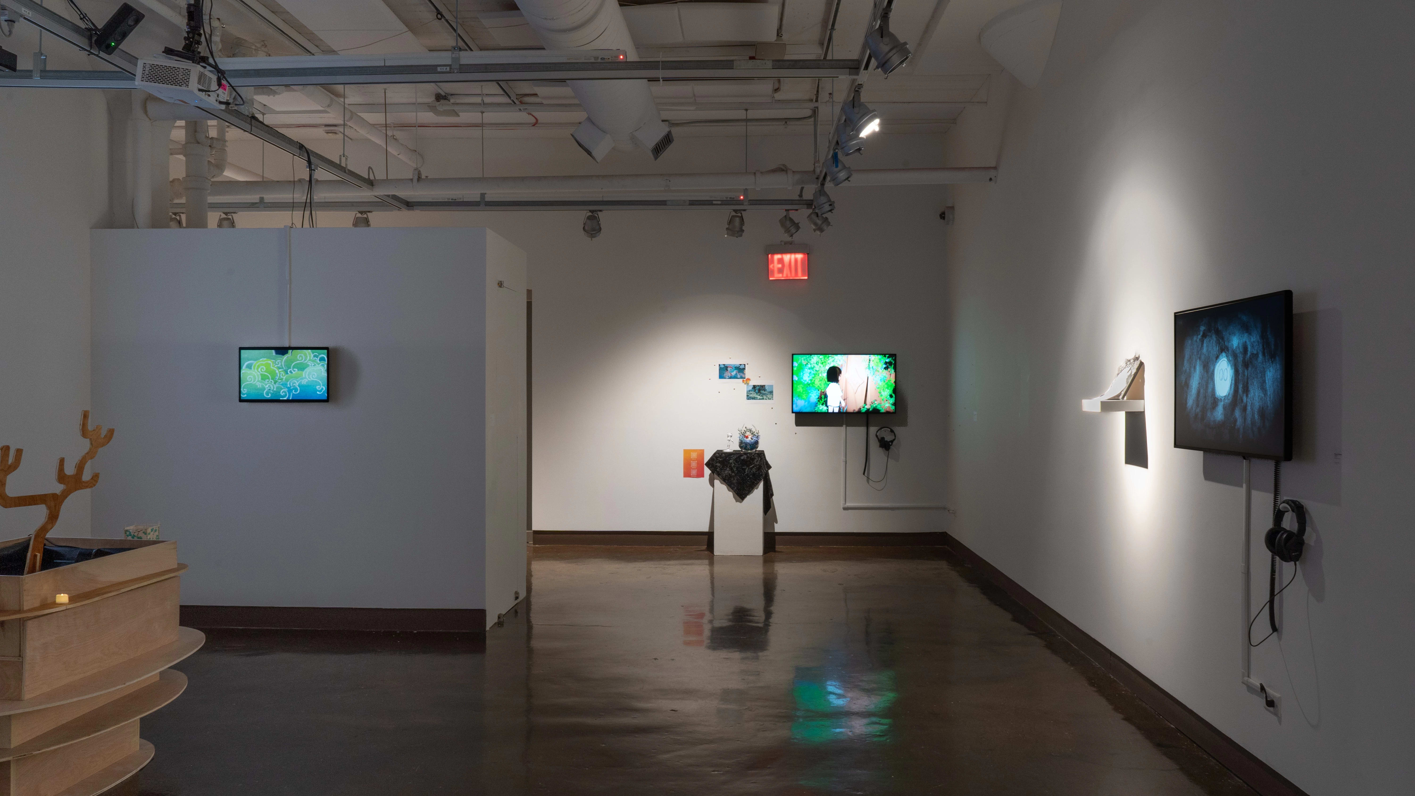 In gallery four, a TV monitor hangs on the wall straight ahead, playing an animated video. Next to it, prints are pinned on the wall, and a pedestal holds a small sculpture. To the left, a large wooden sculpture on the floor is visible in the gallery’s alcove, and to the right, another monitor hangs beside a small shelf.