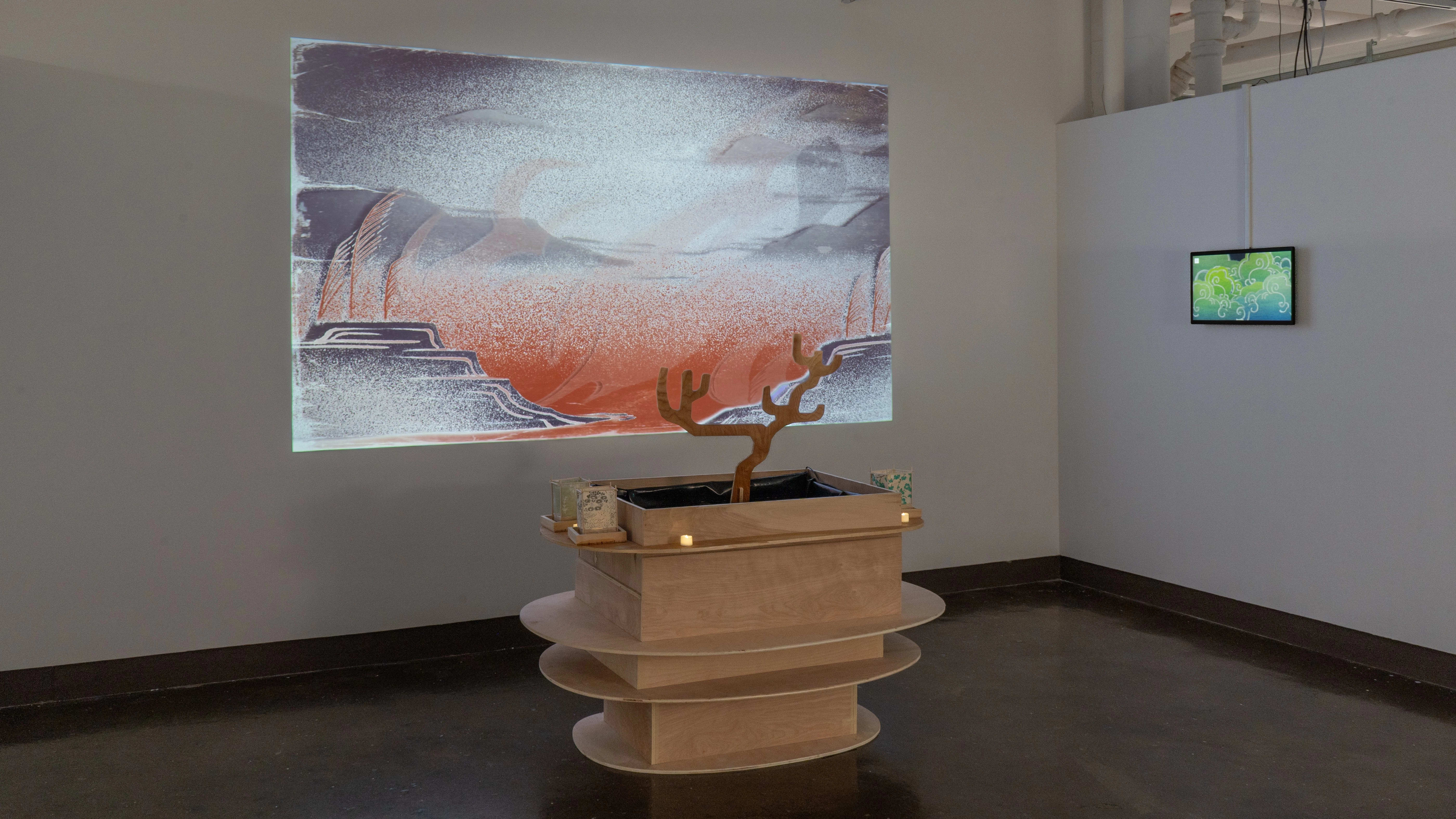 A wooden sculpture of square boxes tiered with oval-shaped shelves sits on the floor in the center of the gallery four alcove. Small lanterns and votive candles sit on the shelf around the top of the sculpture, and an illustrated landscape is projected on the back wall.