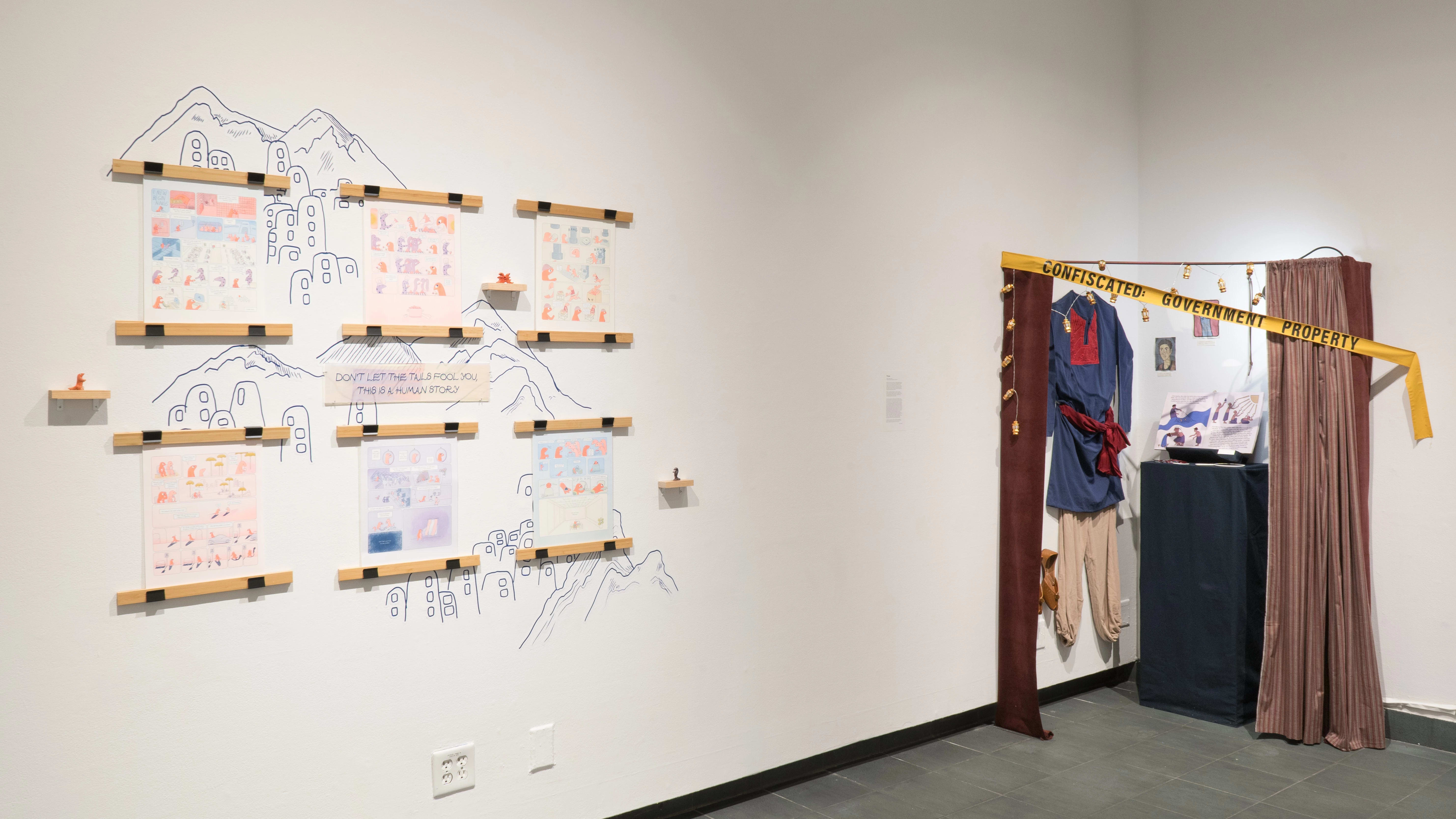 Two works in the exhibition. On the left, a colourful series of comic pages hang on the wall, behind them are mountains represented through blue linework. A plaque reading “Don’t let the tails fool you, this is a human story” sits centred in the piece. To the right of that is a series of items in a corner of the gallery. Clothes and shoes hang on the wall, close to a plinth that has an open book on top of it. The items are behind curtains that are mostly drawn, and yellow tape that reads “Confiscated: Government Property.”