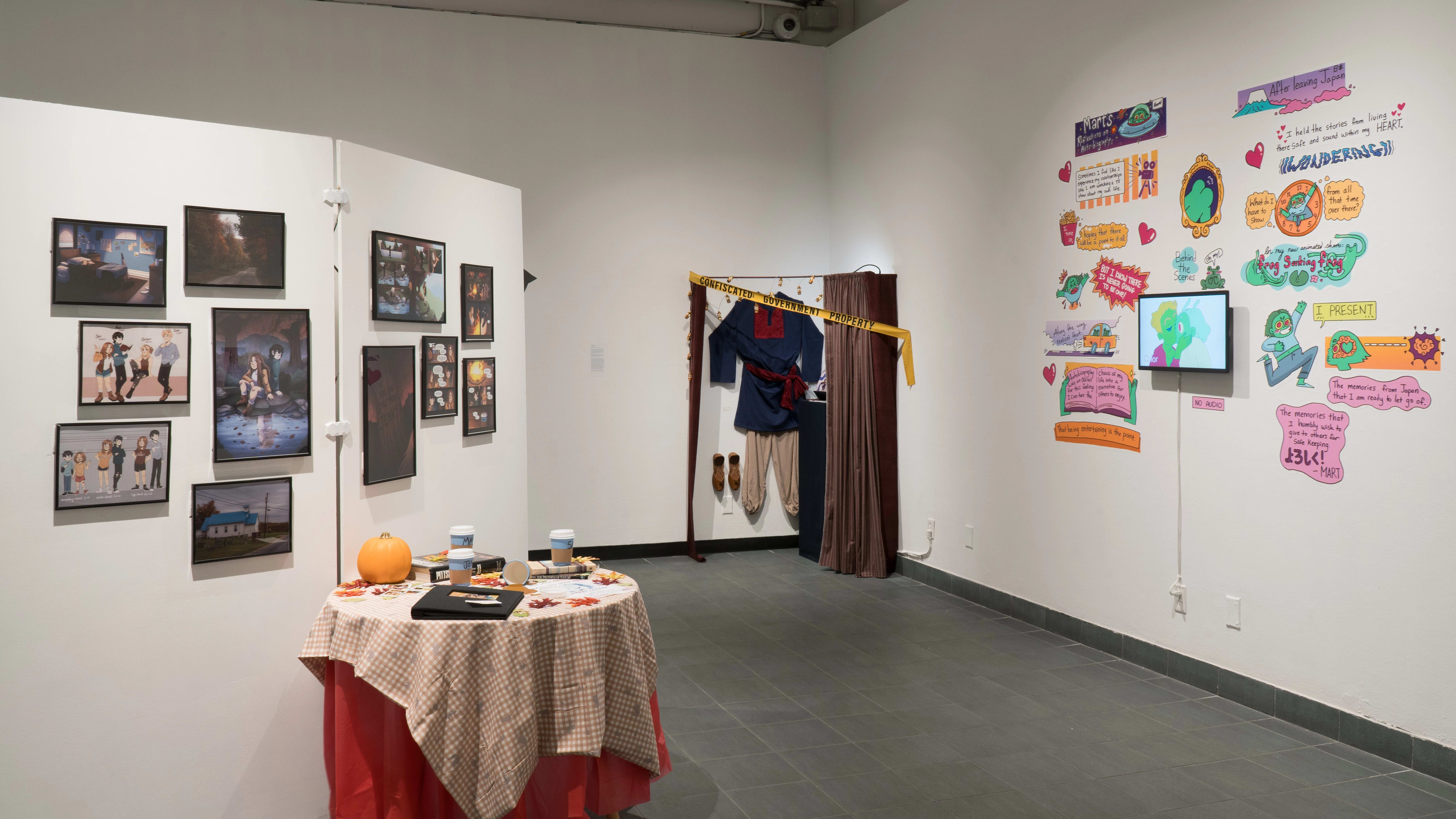 11 framed artworks on a divider in the middle of the room. A small autumn themed table is in front of the works. In the corner of the room is an outfit hung on the wall, partially hidden behind a curtain. To the right is an animation surrounded by graphic stickers, also made by the artist.