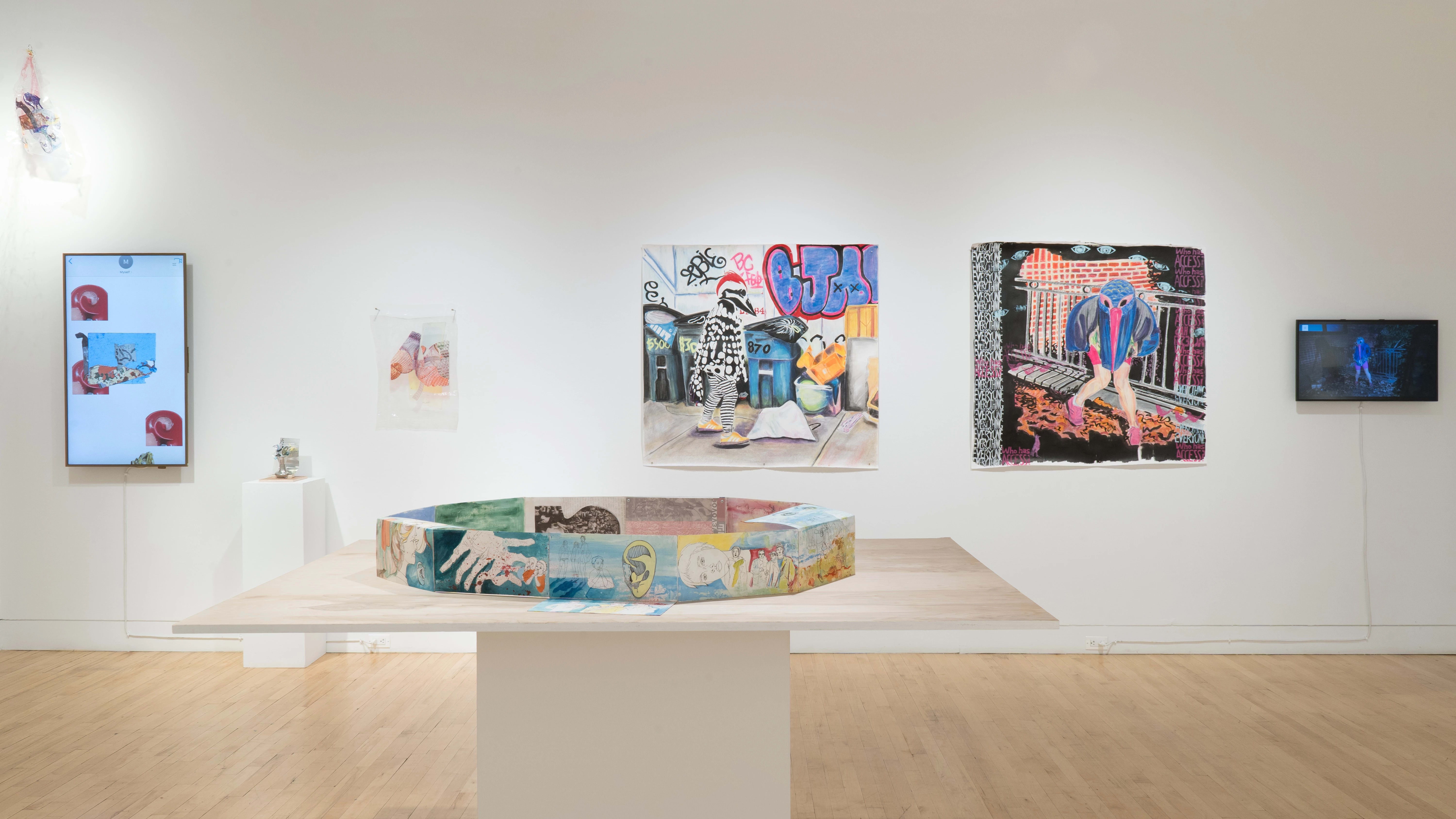 A table in the center of the gallery holds drawings on paper, which are arranged into an oval. On the wall behind the table, there are two monitors, some mixed media works, and two large paintings.
