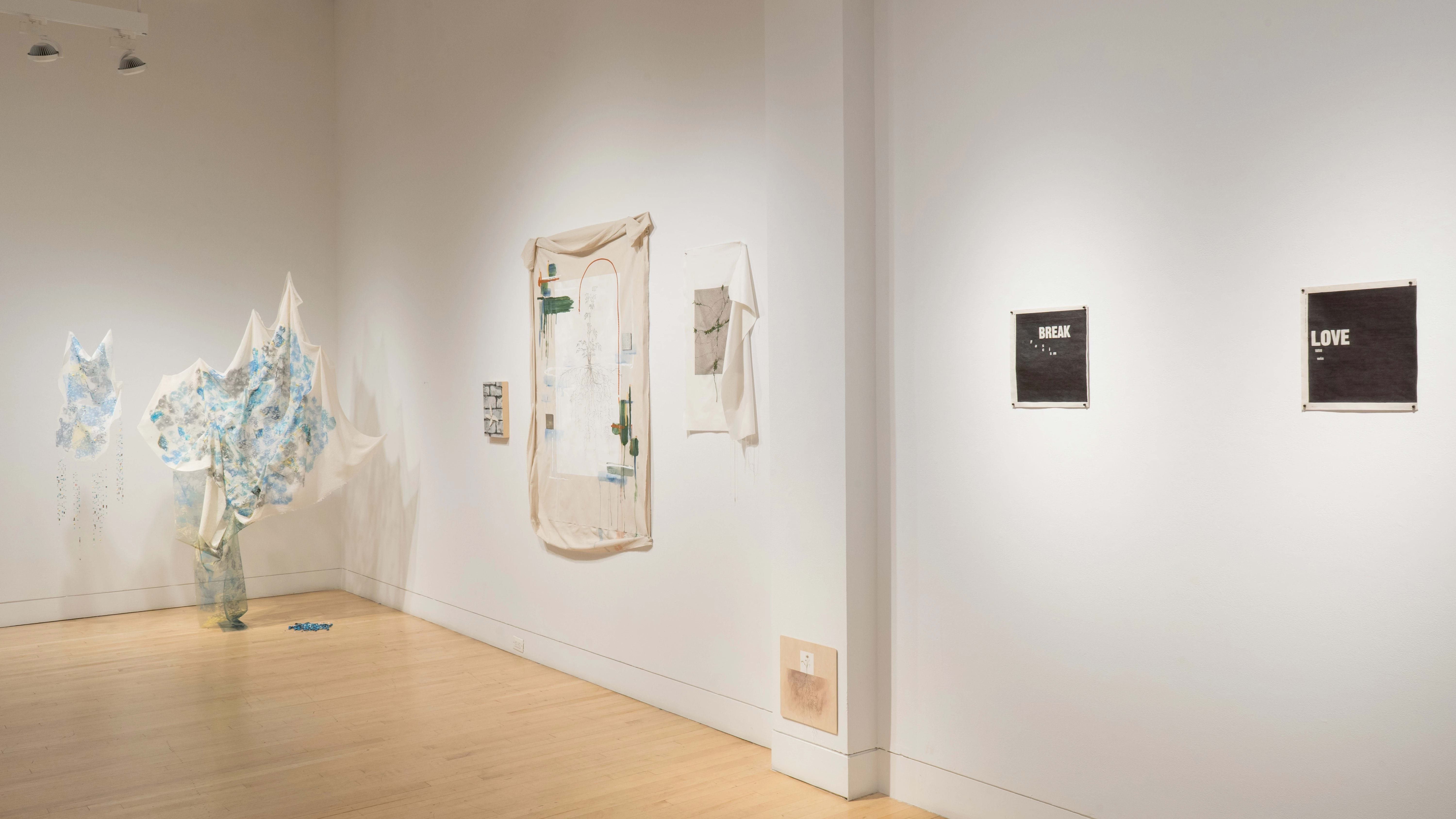 A fabric-based mixed media installation hangs in the corner of the gallery, suspended in the air. To the right, mixed media works on canvas and wood panel hang on the wall. Further, two black prints, one featuring the word “BREAK” and one featuring the word “LOVE,” hang beside one another.