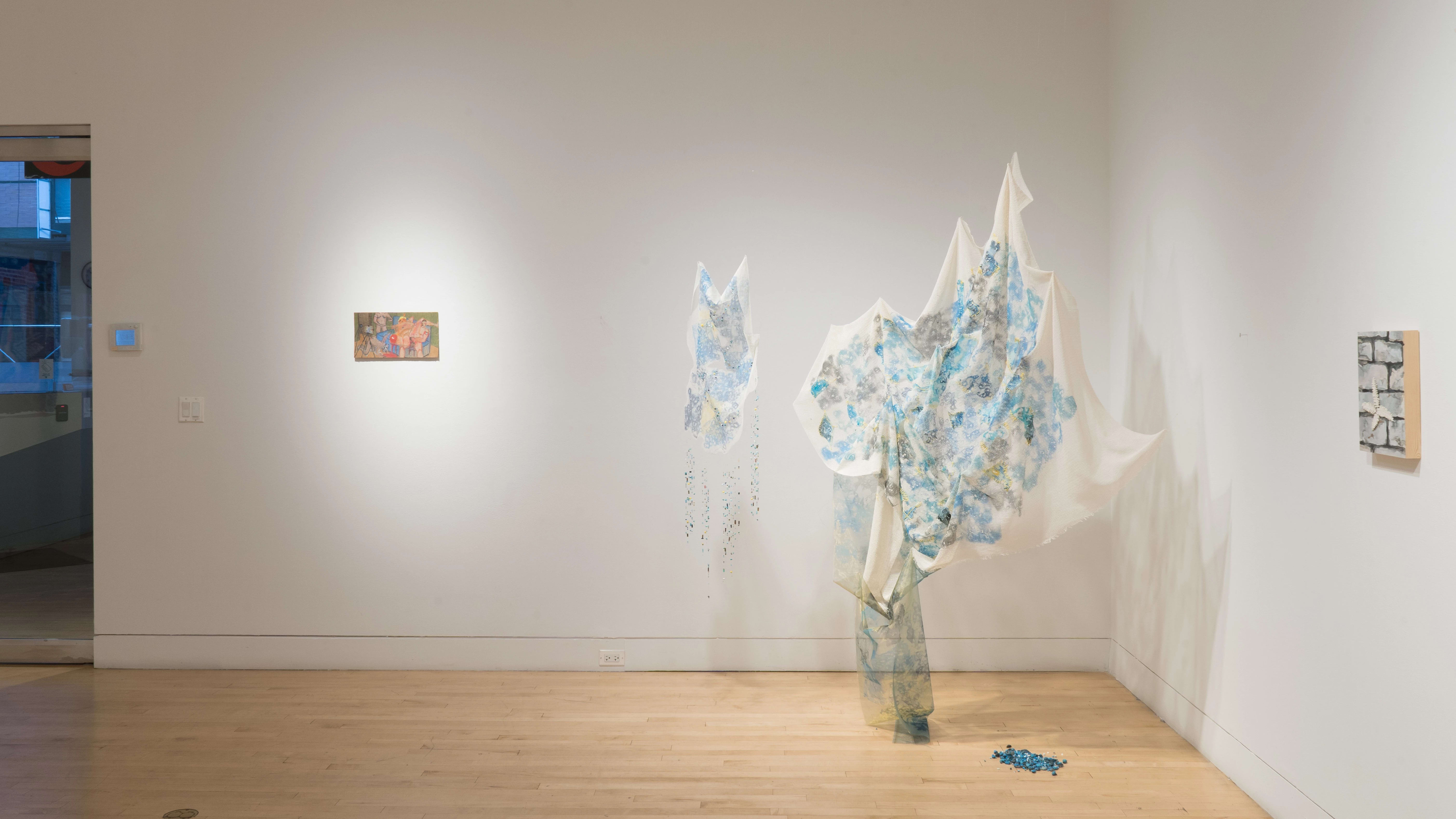 The glass door of the gallery is visible on the left, while a fabric-based mixed media installation hangs in the corner to the right. In the center, a small, horizontal painting hangs on the wall.