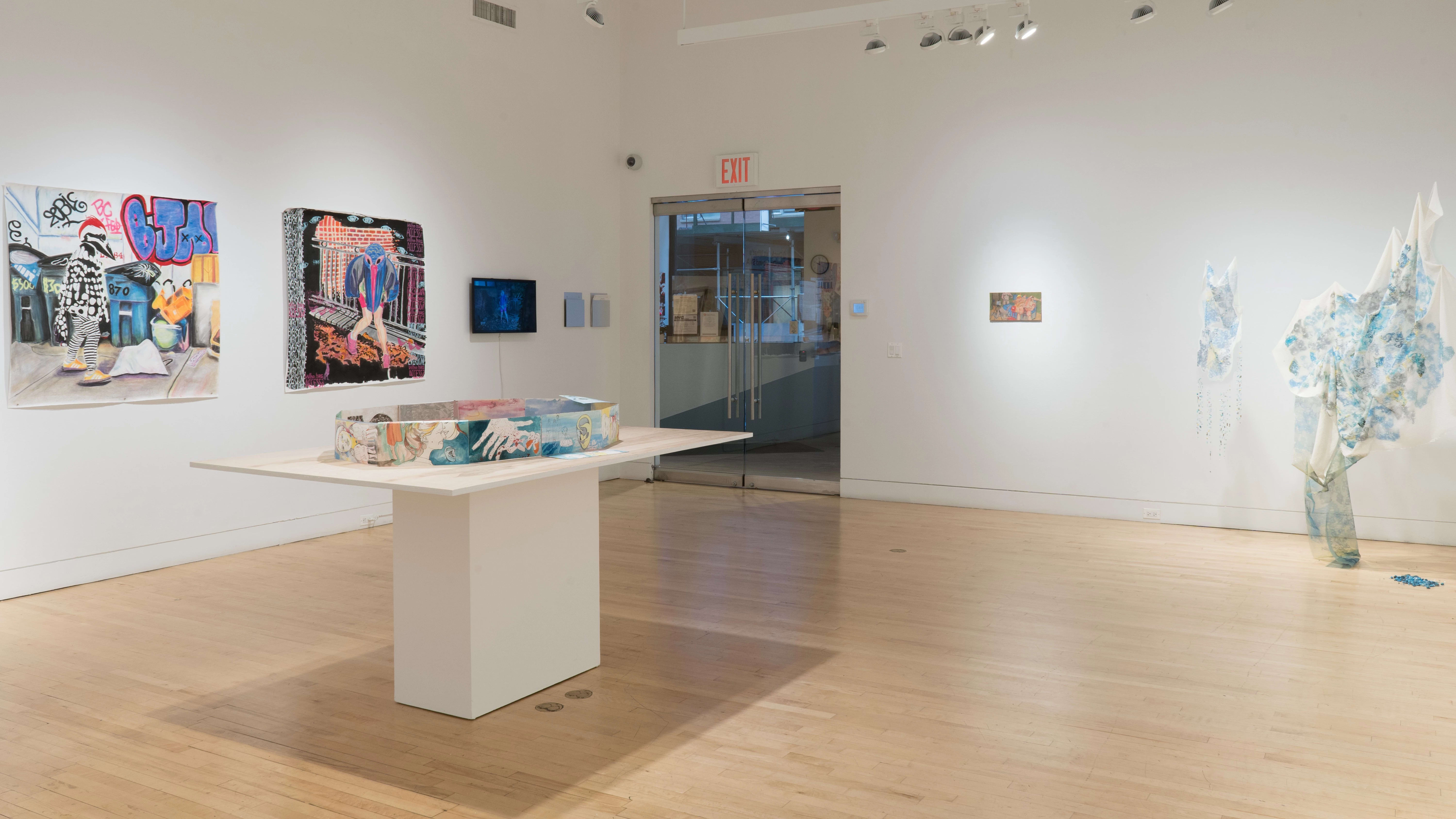 Drawings on paper are arranged in an oval shape on a table in the center of the gallery. Behind it, two large paintings of humans with bird heads hang beside a small monitor. To the right of the table, a small painting hangs next to a fabric-based mixed media installation.
