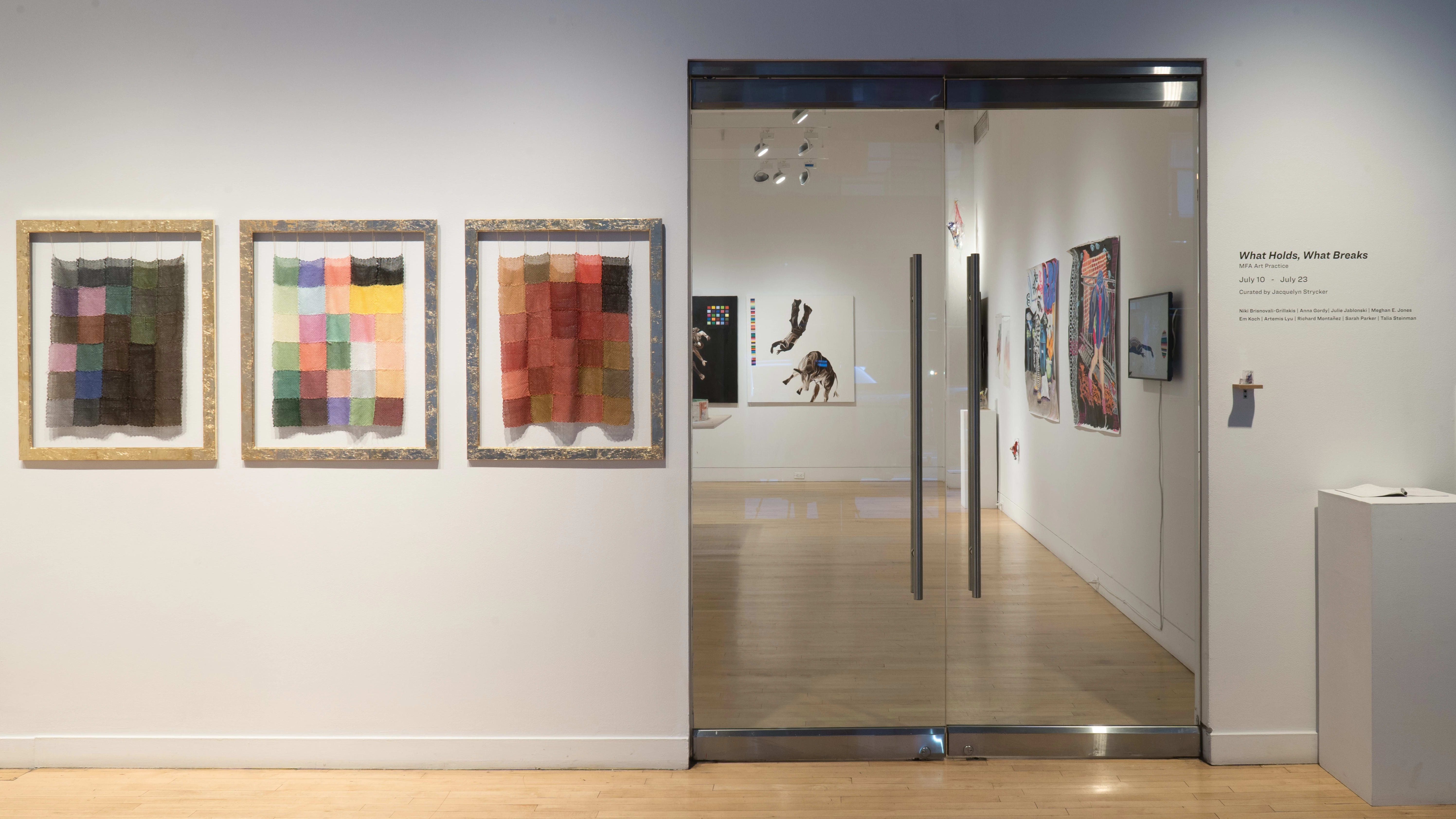 Looking at the gallery from outside the glass doors, some of the work inside is visible. To the right of the door, a very tiny piece is on a small wooden shelf beneath the exhibition title “What Holds, What Breaks.” To the left, three frames hold knit panels with squares of various colors.