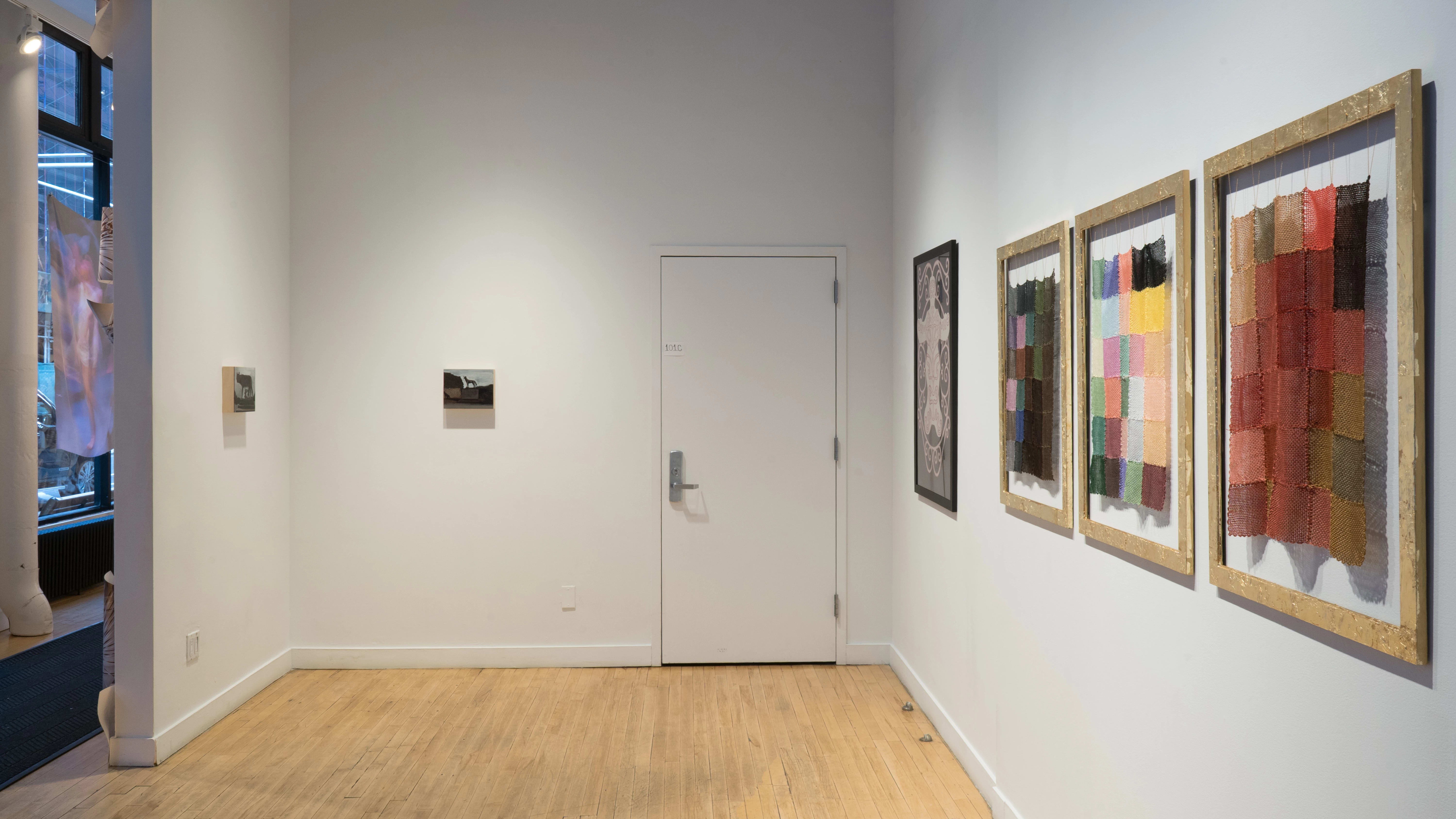 In the alcove outside the gallery, three frames hung on the wall hold knit panels with squares of various colors. Elsewhere in the alcove are two small paintings on wood panel, and one large abstract drawing.
