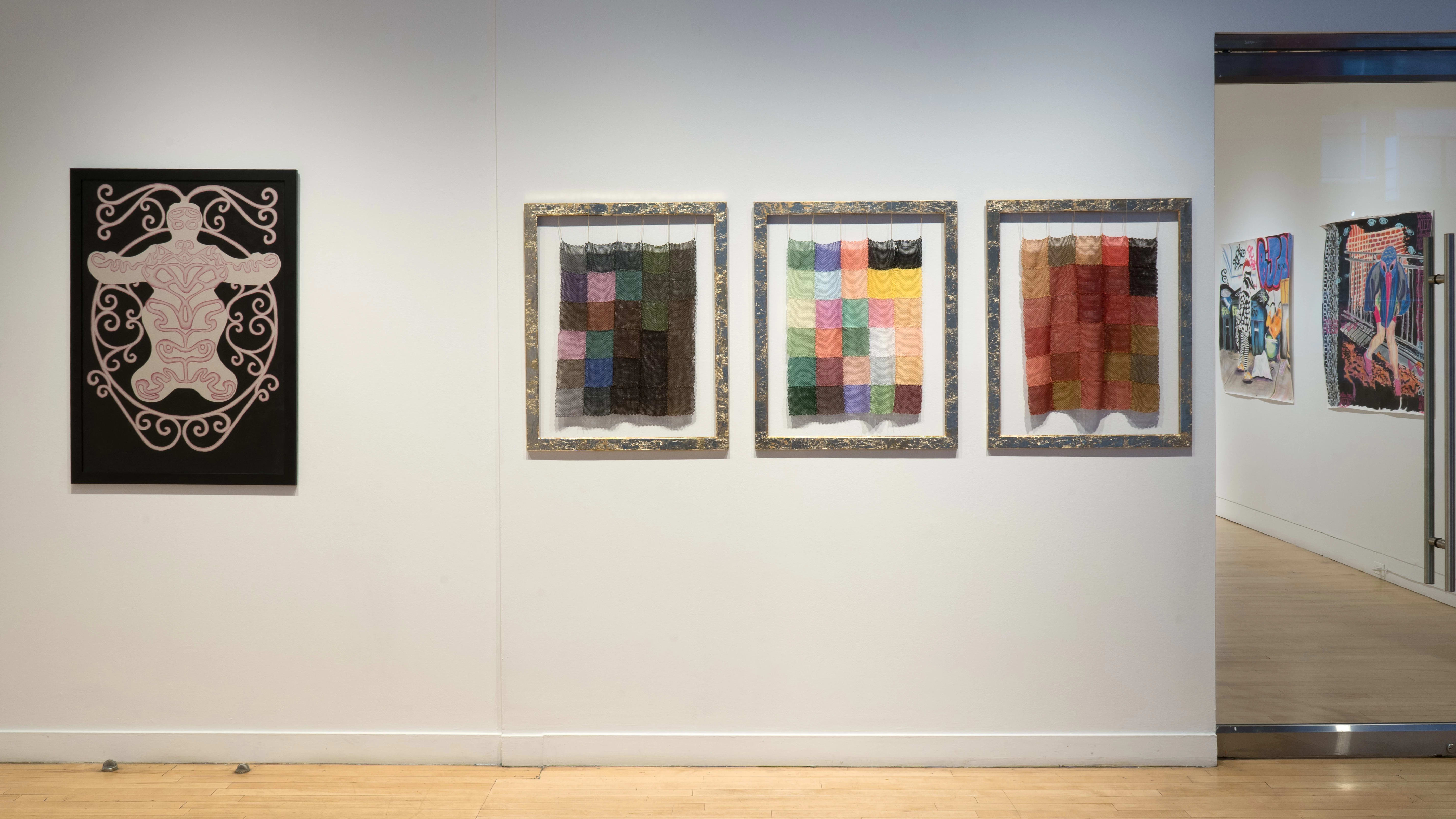 Looking at the gallery from outside the glass doors, some of the work inside is visible. To the left, three frames hold knit panels with squares of various colors. Beyond them, a drawing in black, pink, and neutral depicts a body-like shape with a swirling pattern.