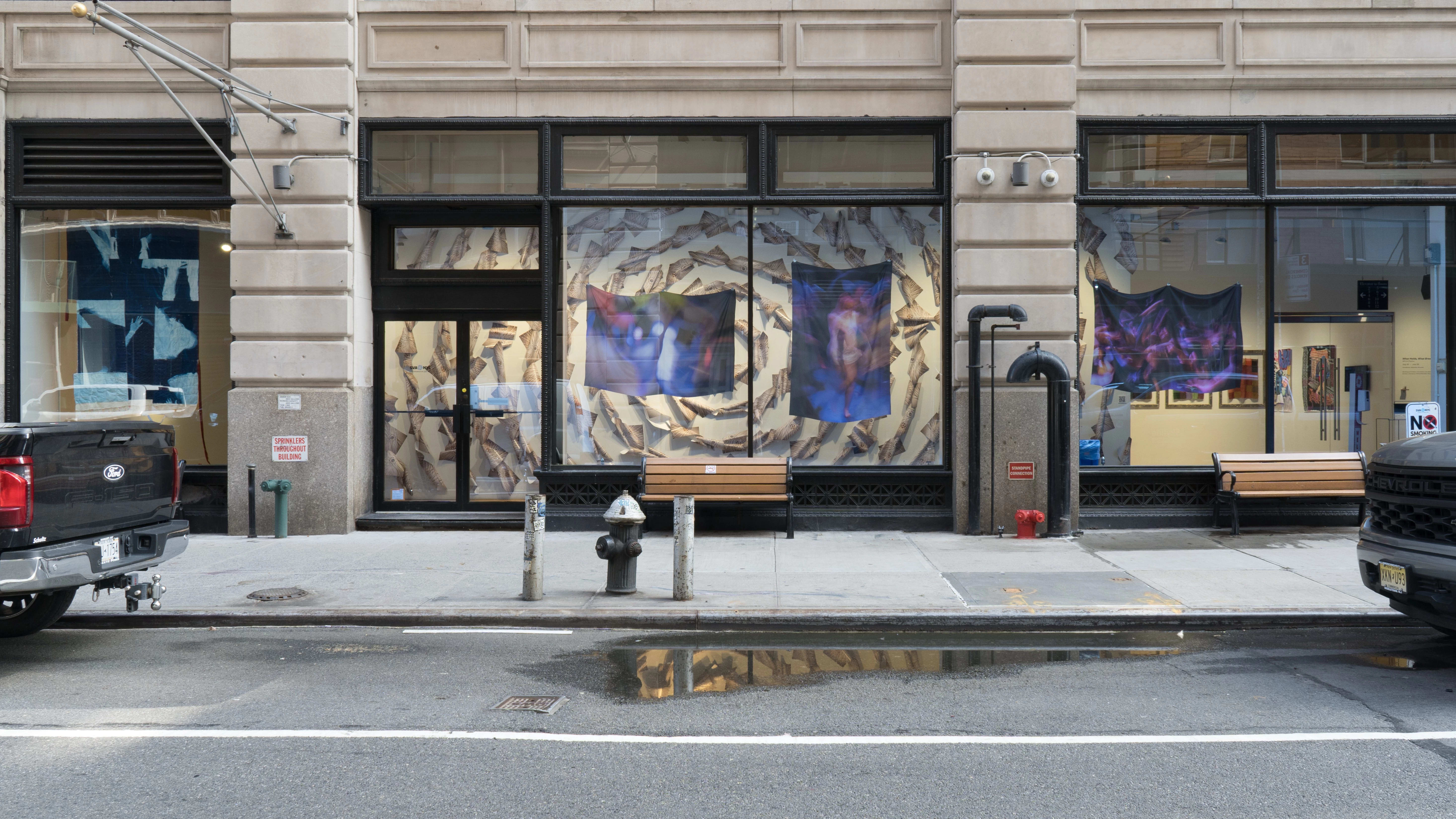 From outside the SVA Flatrion Gallery building, several works on fabric hang in the windows. One appears to be a quilt, while the other three are blurry photos printed on fabric. Behind them, many papers are arranged into circles radiating from the center point of the wall.