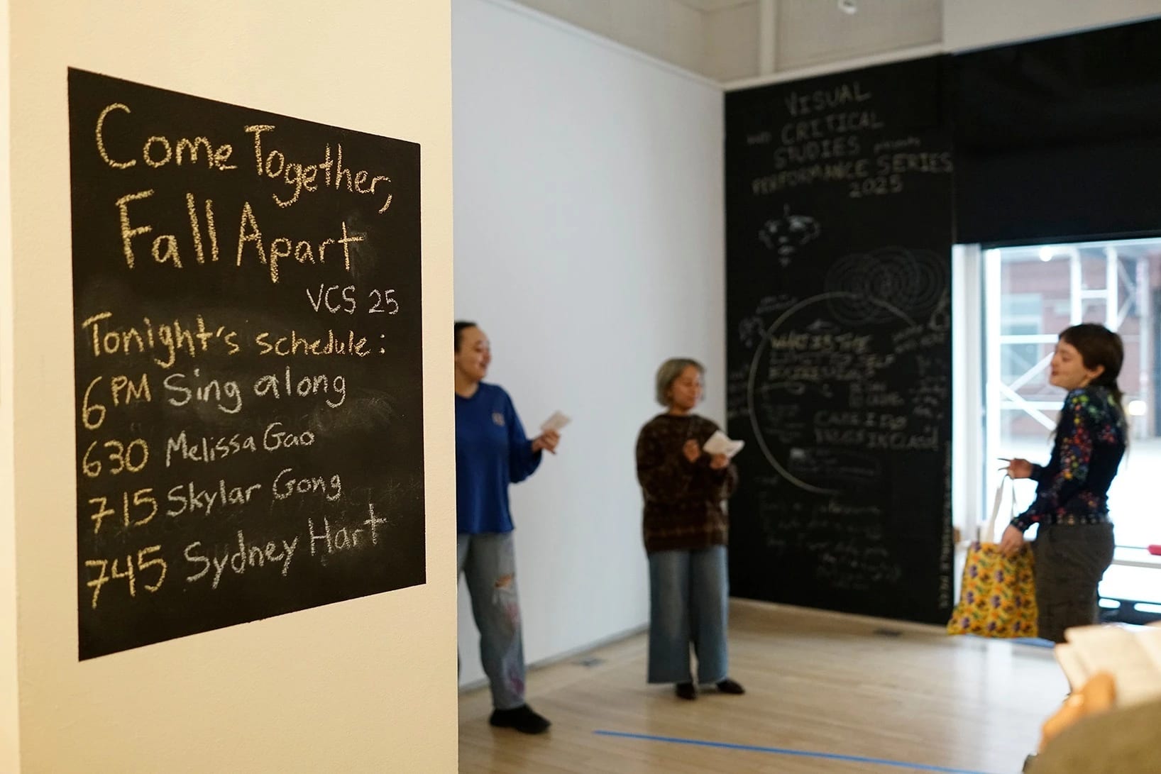 Shot of the sing-a-long performance in teh Flatiron Project Space from the entrance, with a sign of the day's performance schedule on the left.