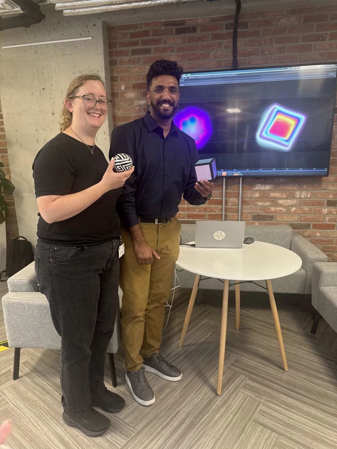 Two people pose in a lounge holding up toys that correspond to a TV behind them