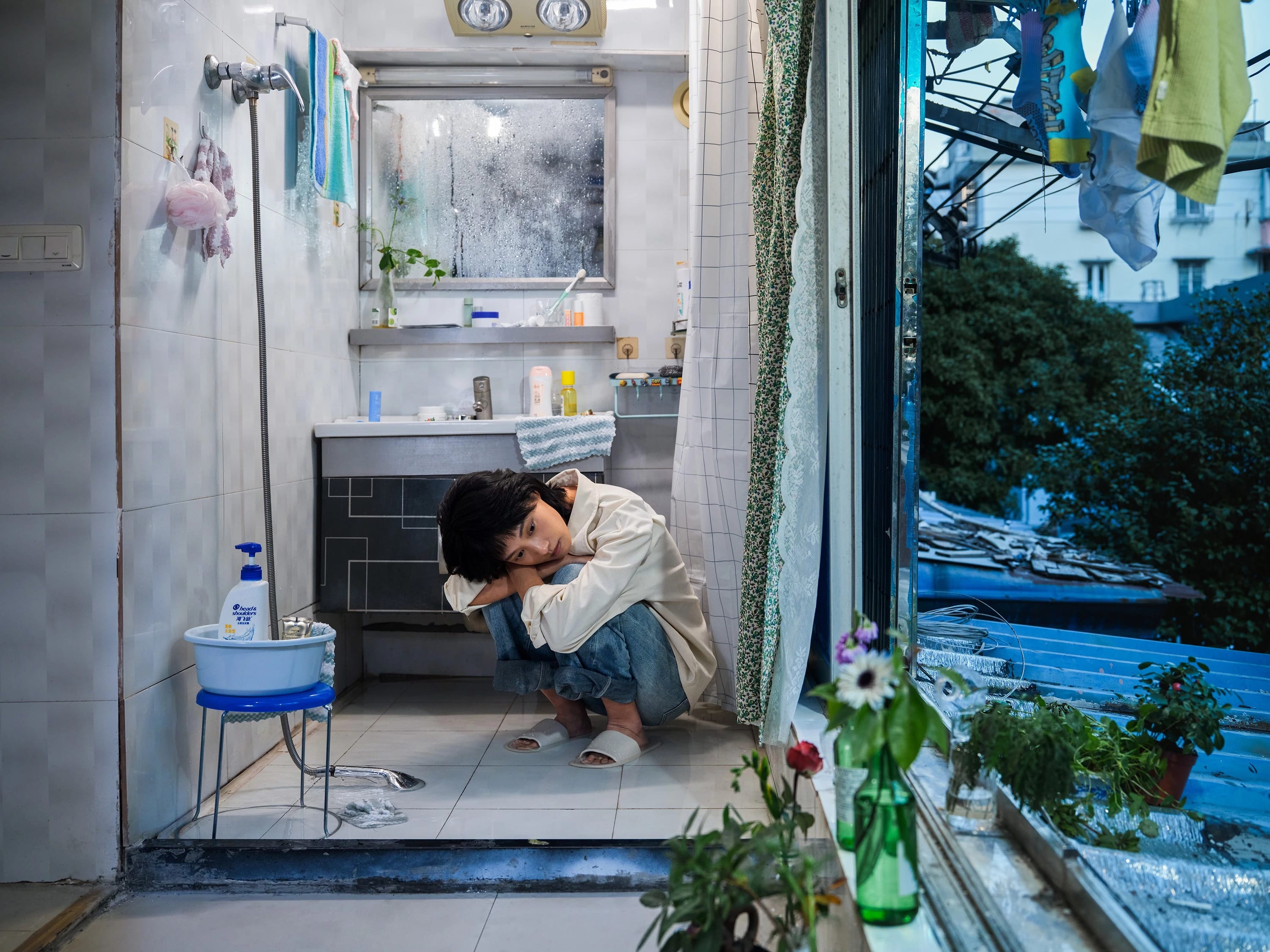 A color photography by Shaohan Fang entitled En Route. In a dim bathroom at dusk, the girl crouches on a wet floor, staring at the window. Water pools around her feet, she appears visibly overwhelmed.