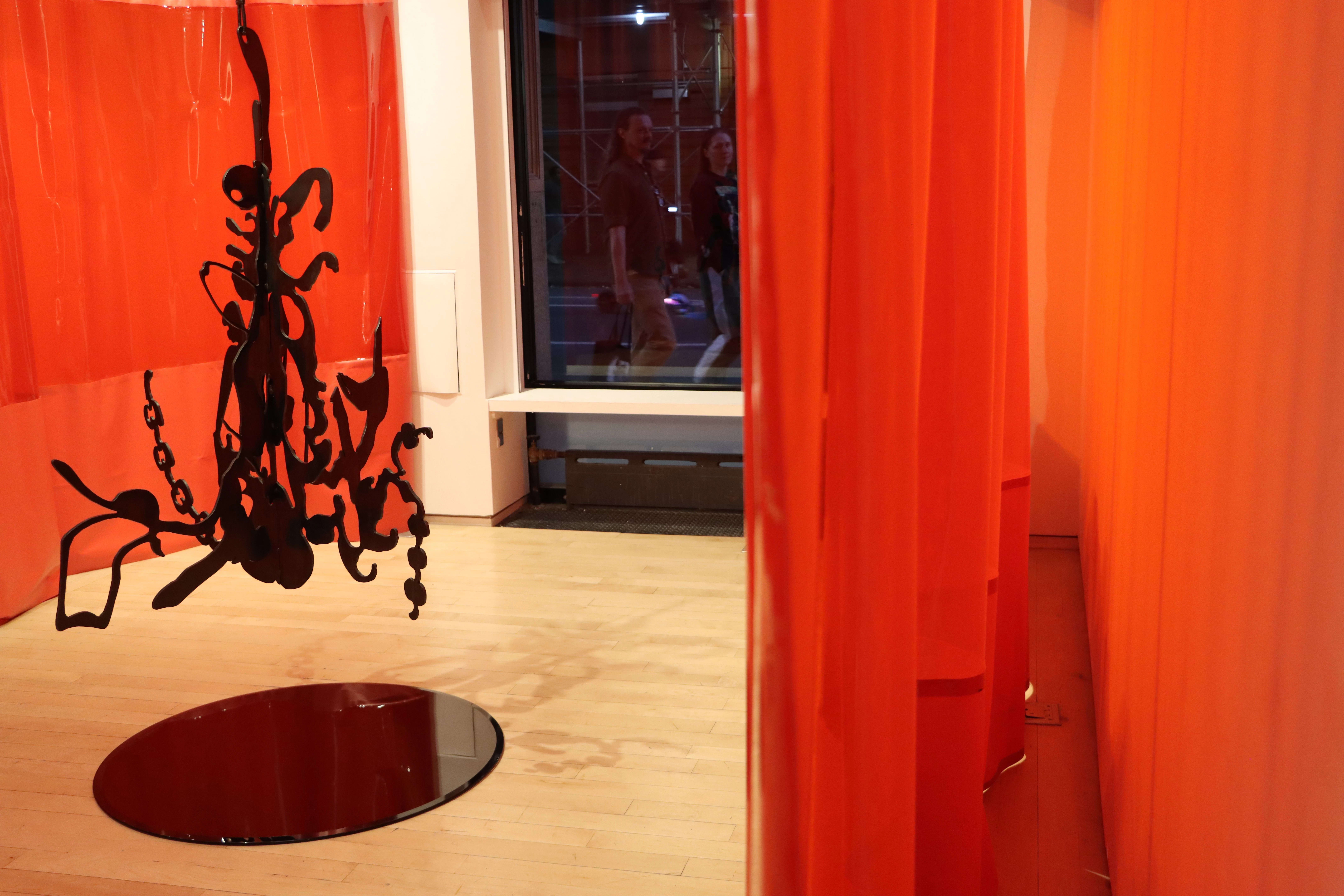 nstallation shot showing a black plexiglass organic shape hanging from the middle of the room surrounded by a red curtain and a black plexi circle immediately beneath it.