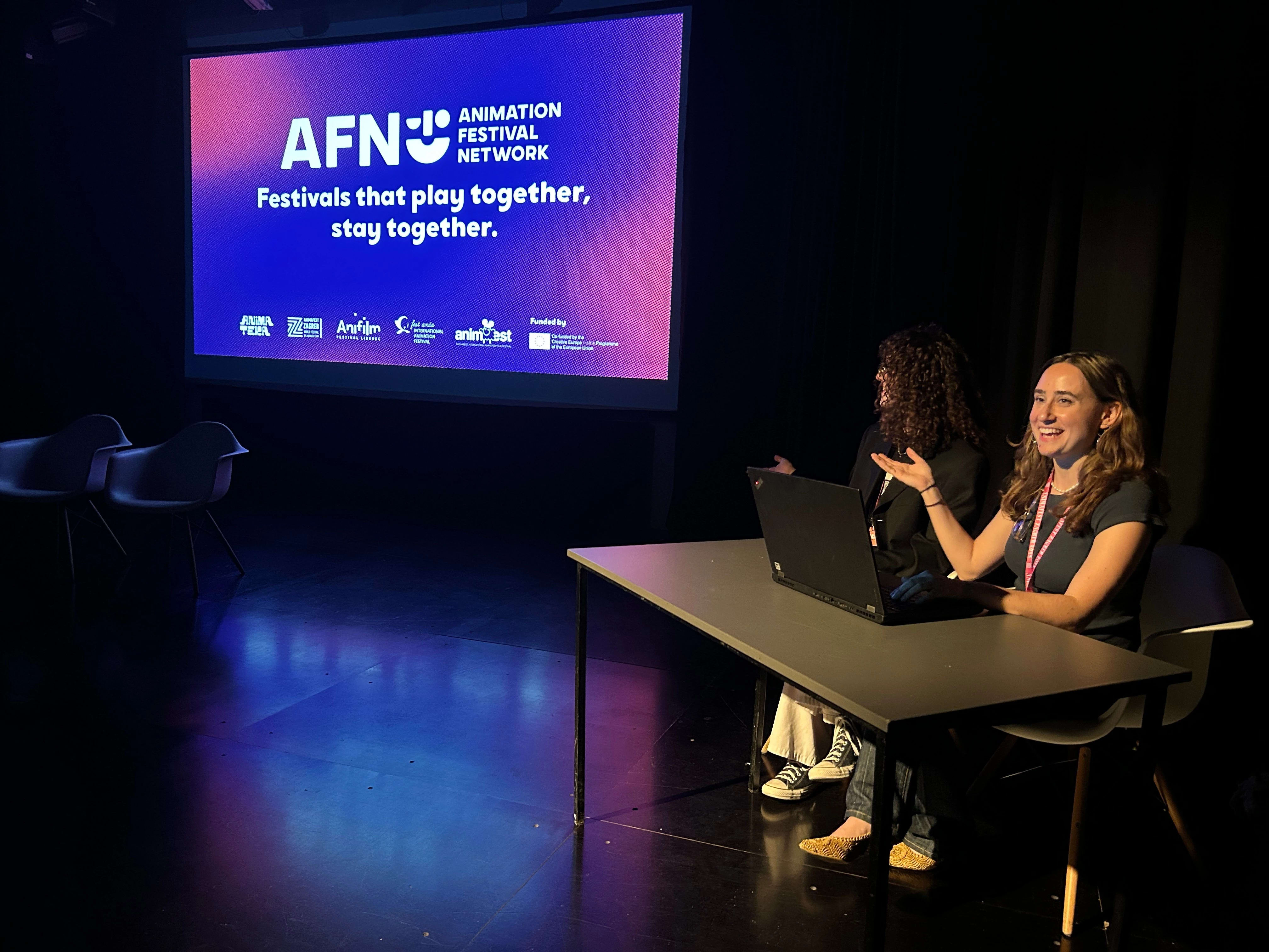 Two people sit at a desk presenting and gesturing toward a projected screen