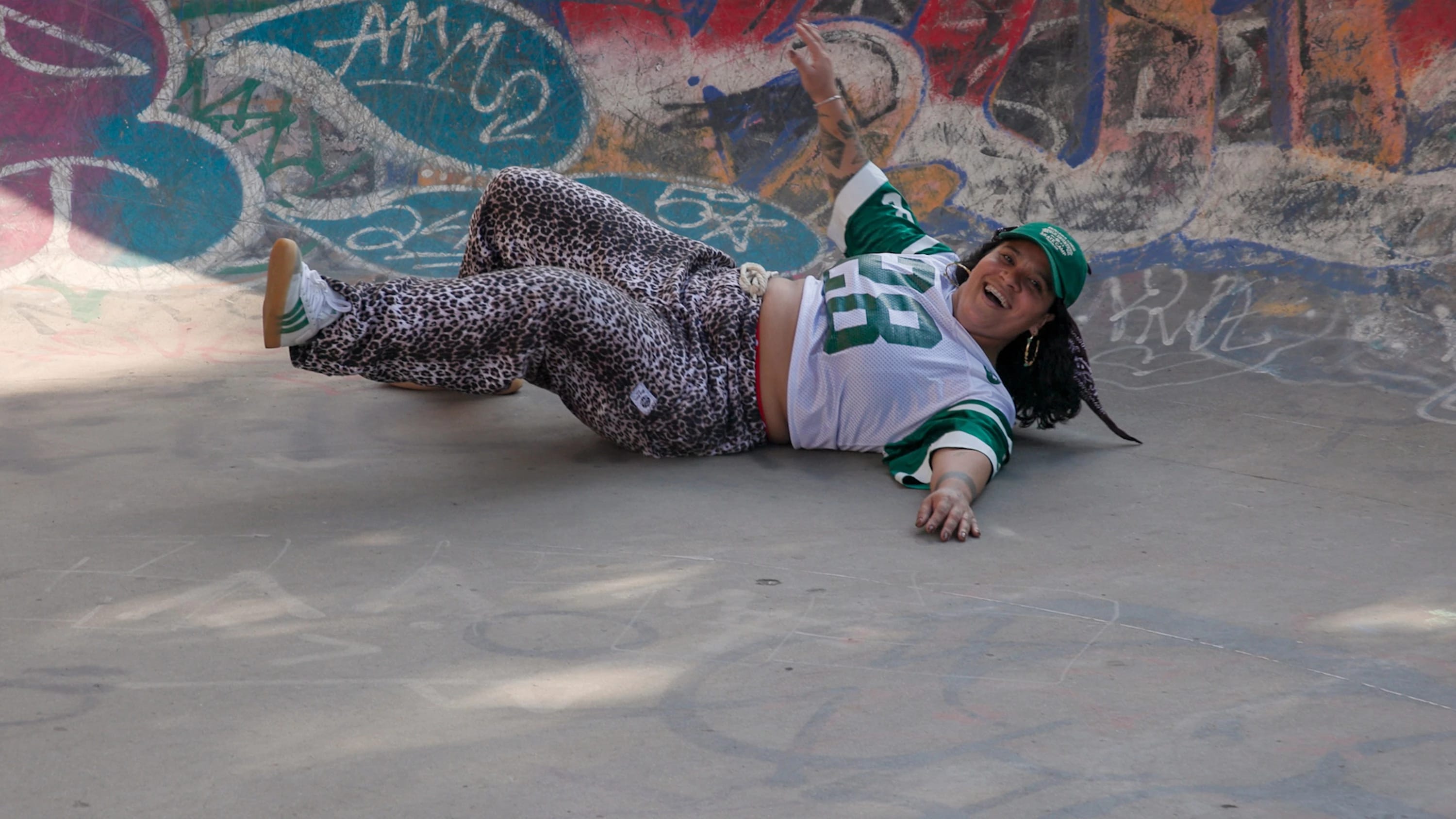 Color still from FREE SKATE (dir. Lia-Alexi Manfredi, 2025). A woman in leopard-print pants and a green and white sports jersey lies in a graffiti-covered skate bowl, laughing after a wall with one arm raised.