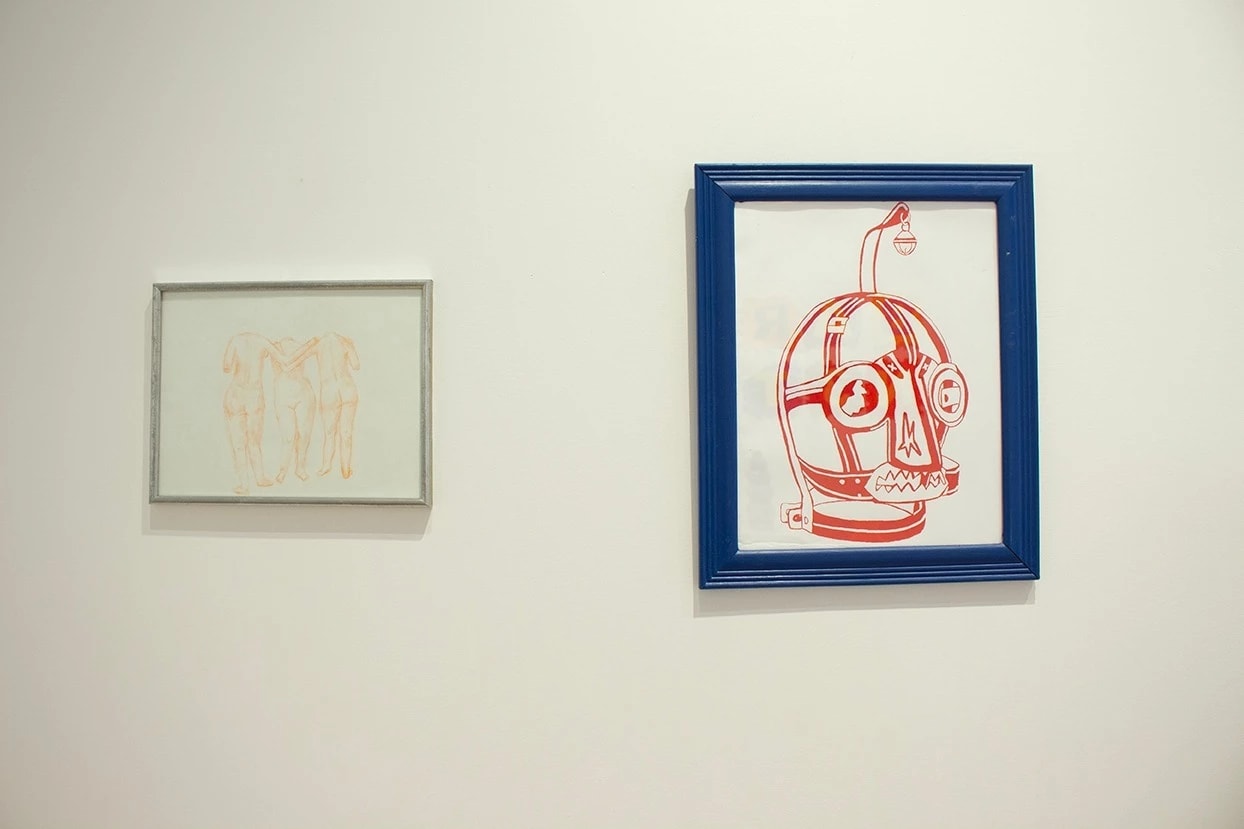 Installation shot of two framed pieces, the one on the left a drawing or nude bodies and the one of the right a print of a leather mask.
