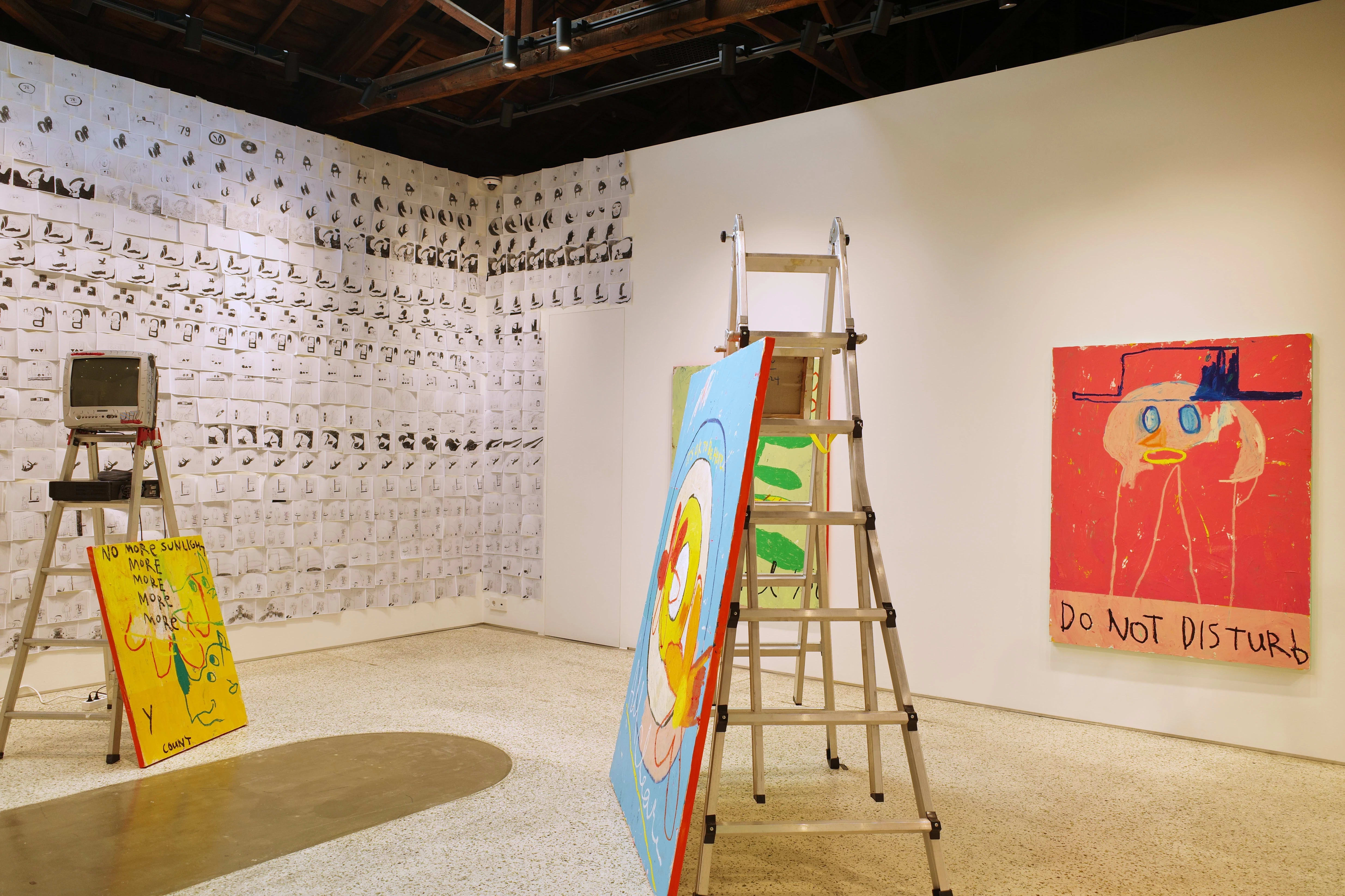 Gallery installation with expressive, cartoon-like paintings and a wall covered in black-and-white sketches. A colorful duck painting leans on a ladder, and another canvas reads 'DO NOT DISTURB' below a pink face in a hat. A vintage TV sits atop a second ladder.