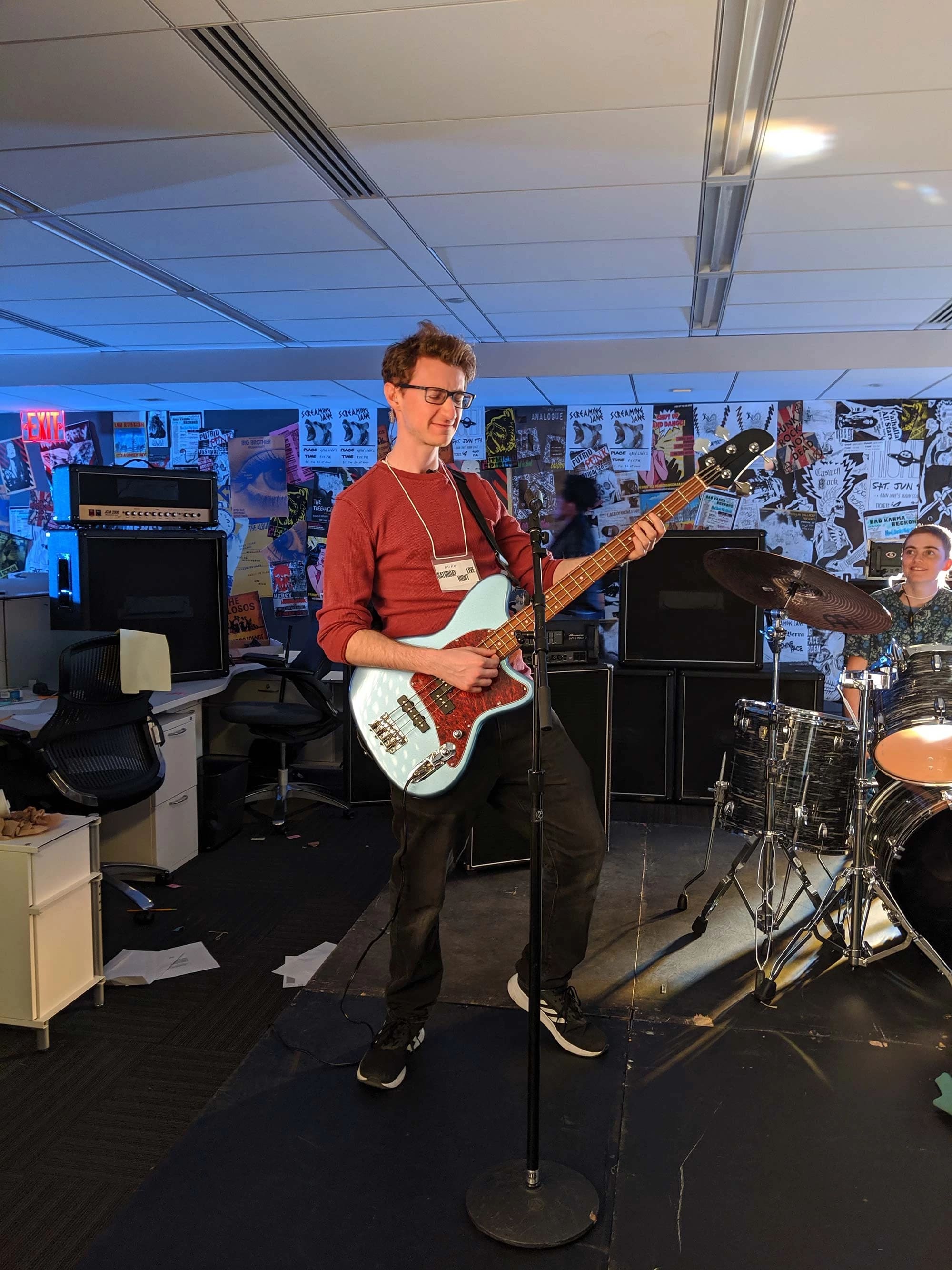 A musician playing a blue bass guitar while standing on stage. Behind him, a drummer is seated, and the background features posters on the walls. The setting appears to be a music space with various instruments and equipment.