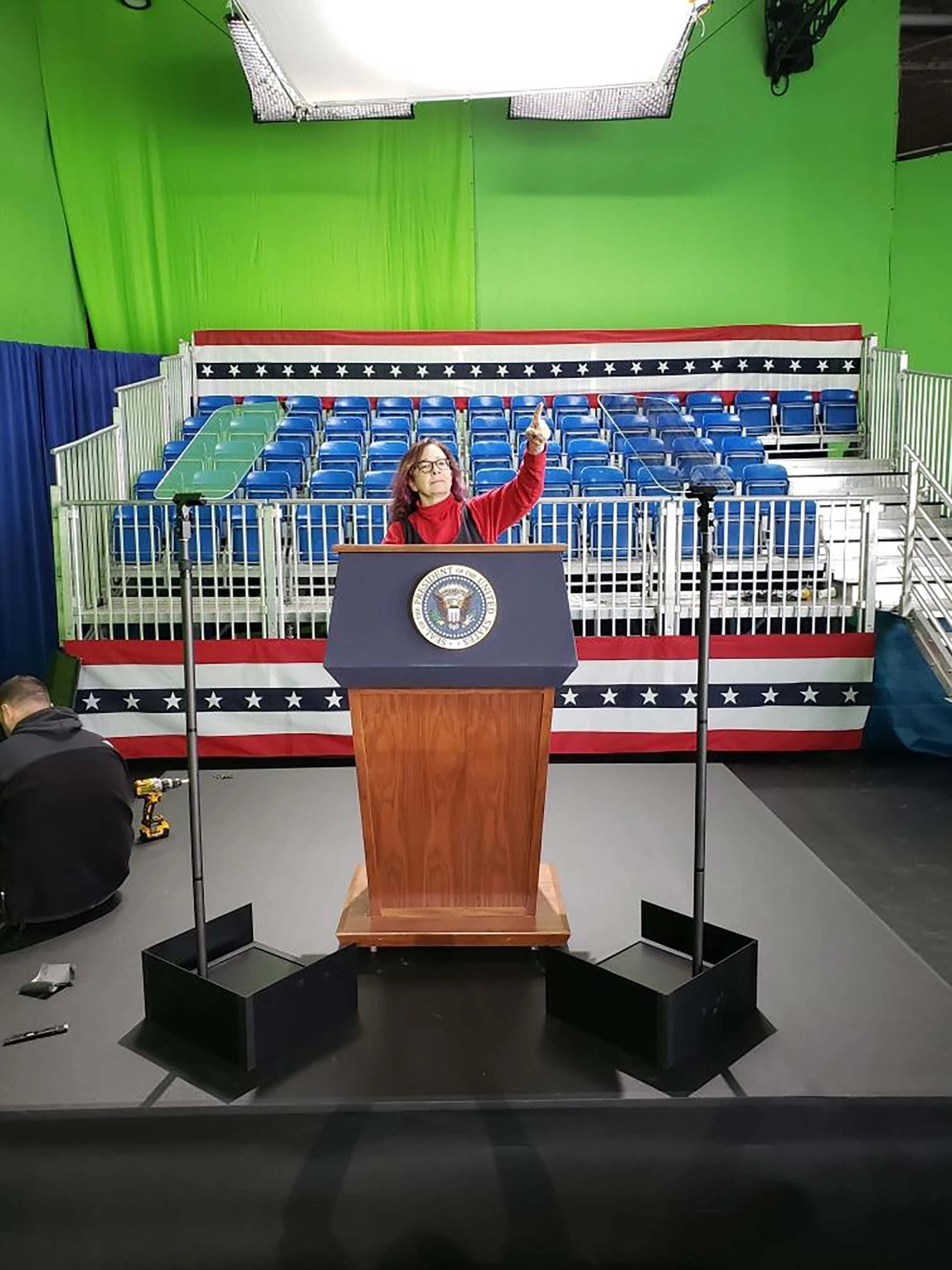 A woman in a red jacket stands at a podium with the presidential seal, facing an empty audience area with blue folding chairs arranged in rows behind her. The backdrop features a red, white, and blue design. Lighting is set up above the podium.