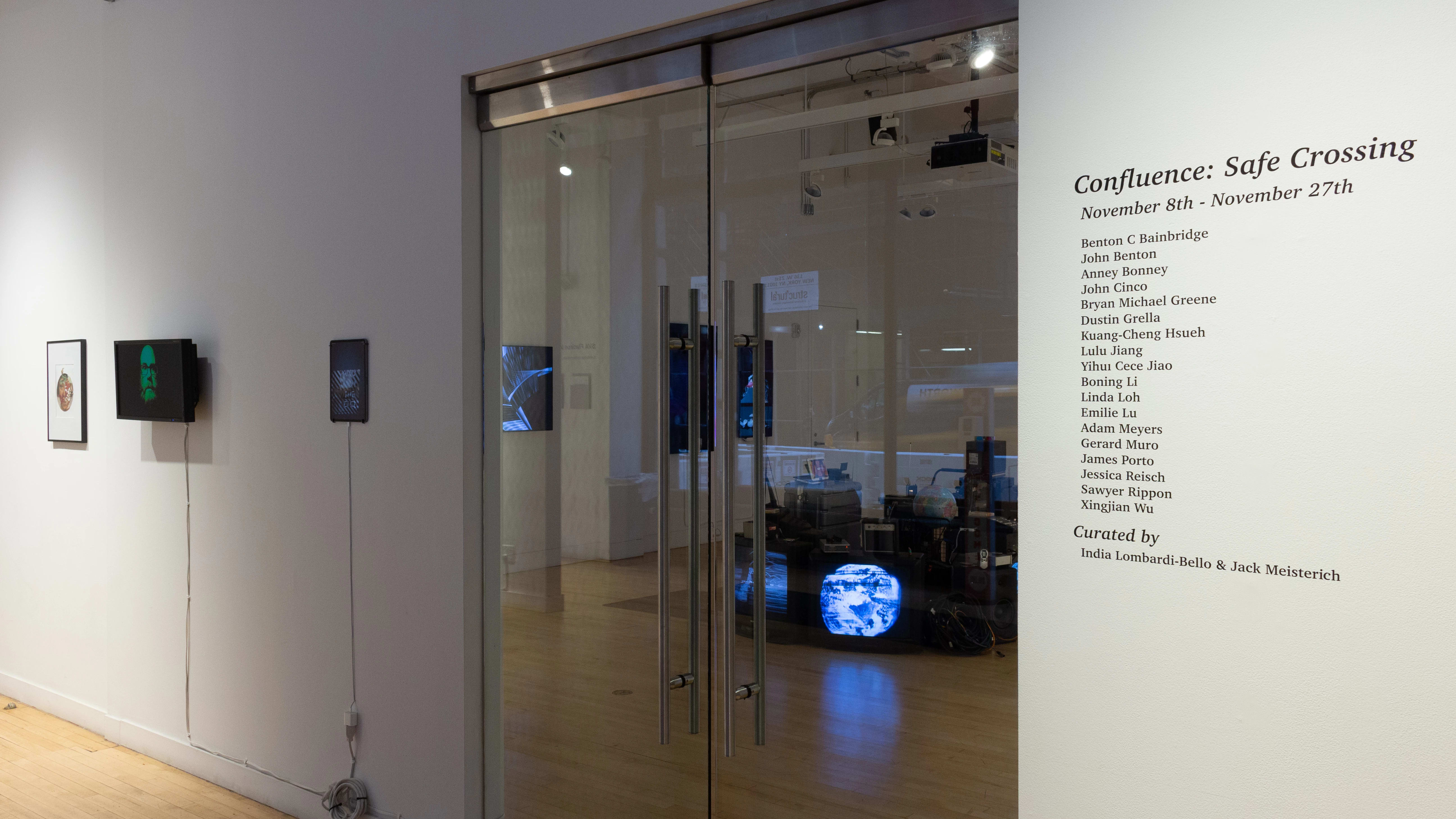 Through the gallery’s glass door, several television monitors and an installation that includes various technological elements are visible. To the right of the door, there is wall vinyl text listing the exhibition’s title, artists, and curators, and to the left of the door, there are more monitors and a framed print.