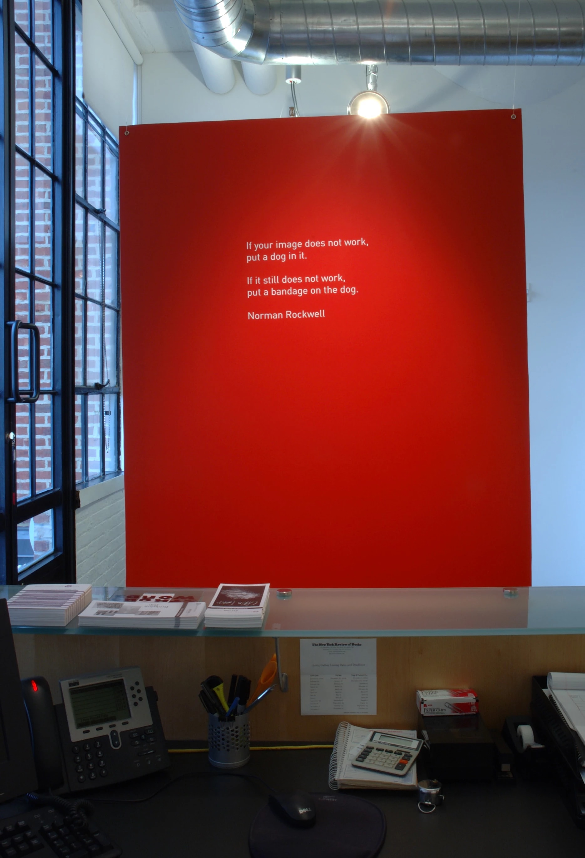 This shows the top of a desk with a large red colored portion of a wall in front of it that has a quote by Norman Rockwell.