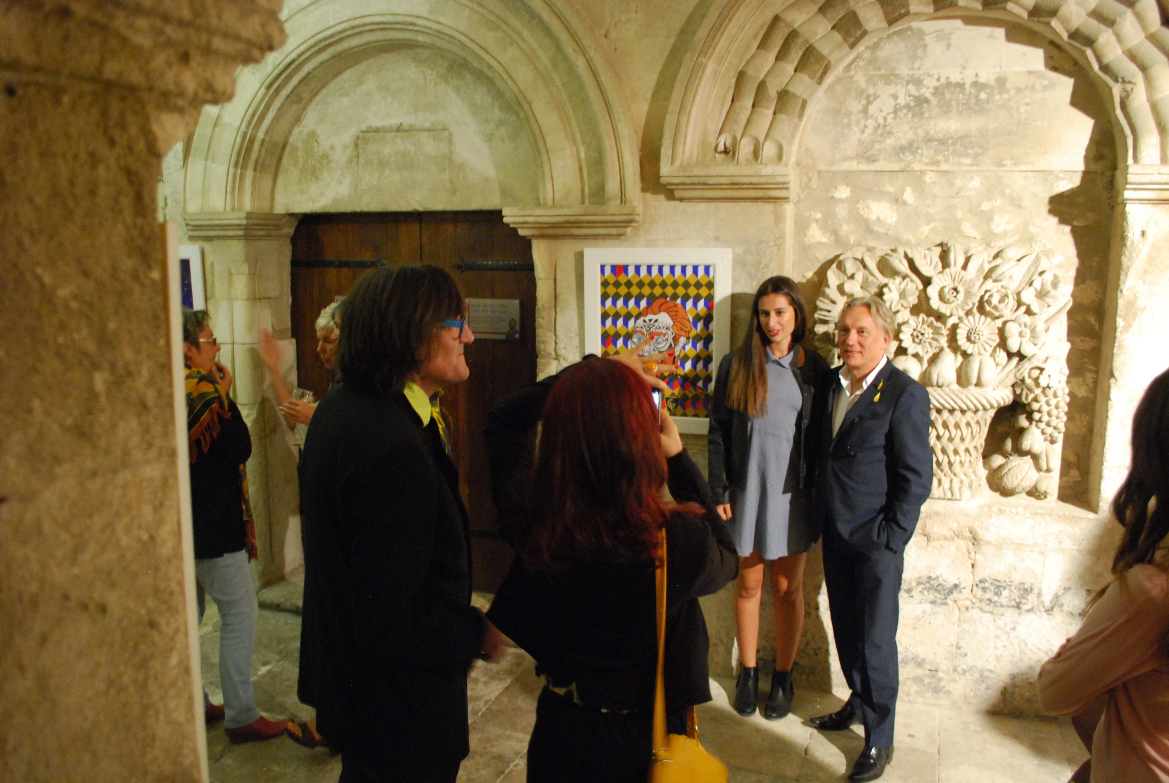A group of people stand in the cloister of Saint-Paul de Mausole during the opening of the exhibition. Beside them is a framed work using blue, yellow, and orange geometric shapes, and bhind them, flowers and fruits are carved into the wall.