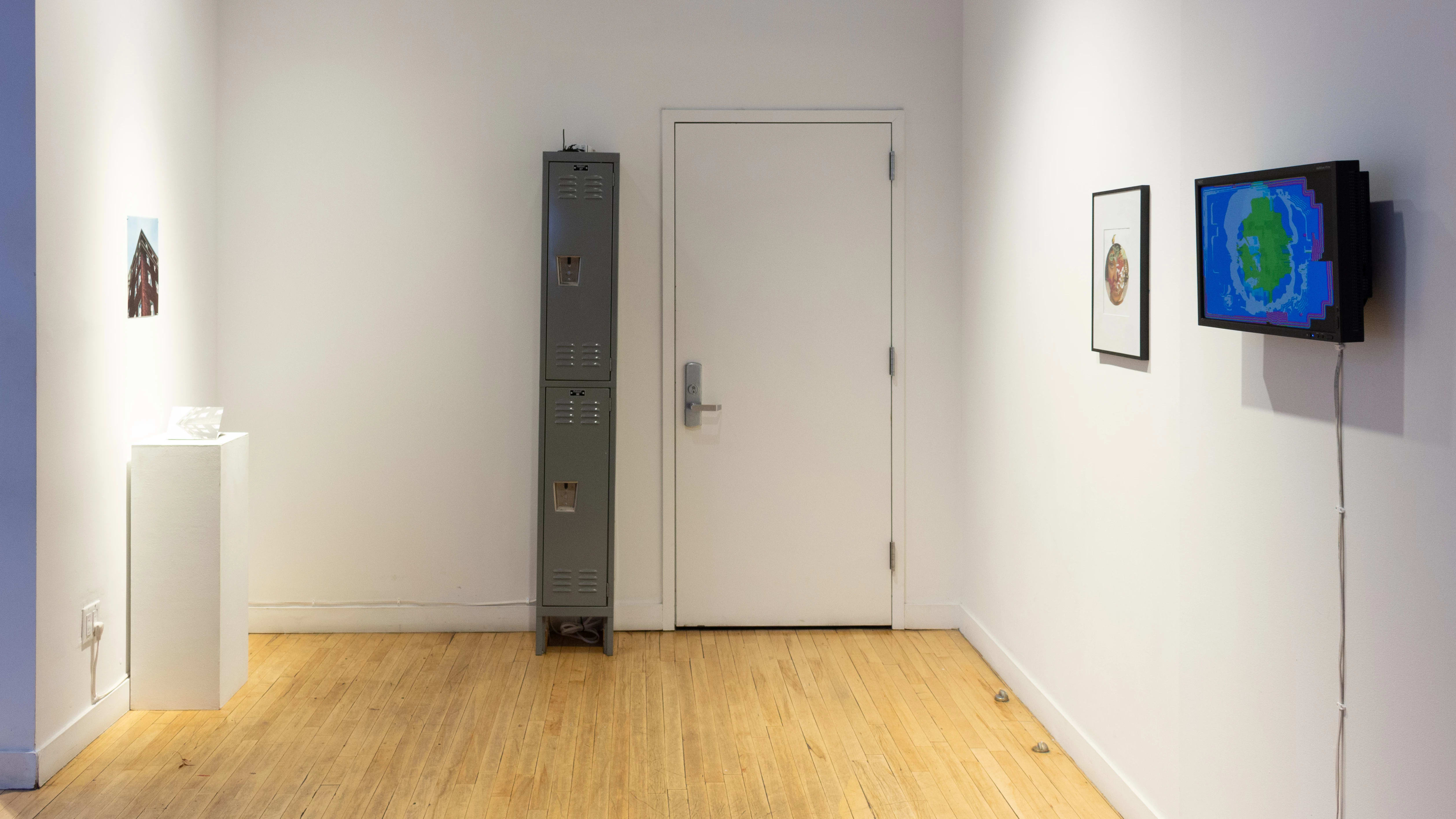  In one corner of the gallery, a narrow vertical locker stands next to a closed door. To the left, a print is pinned on the wall, and to the right, a framed print and a tv monitor hang beside each other.