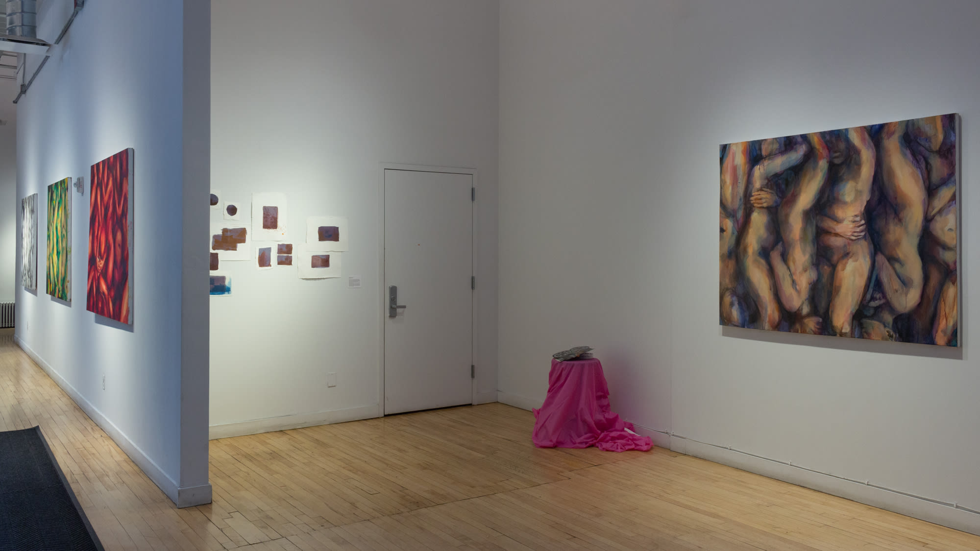 Photograph with view of paintings hung in hallway to far left, then in an alcove to the right are multiple small pieces on paper, a low pink table with an object on it, and to the farthest right is a painting of abstract fleshy bodies.