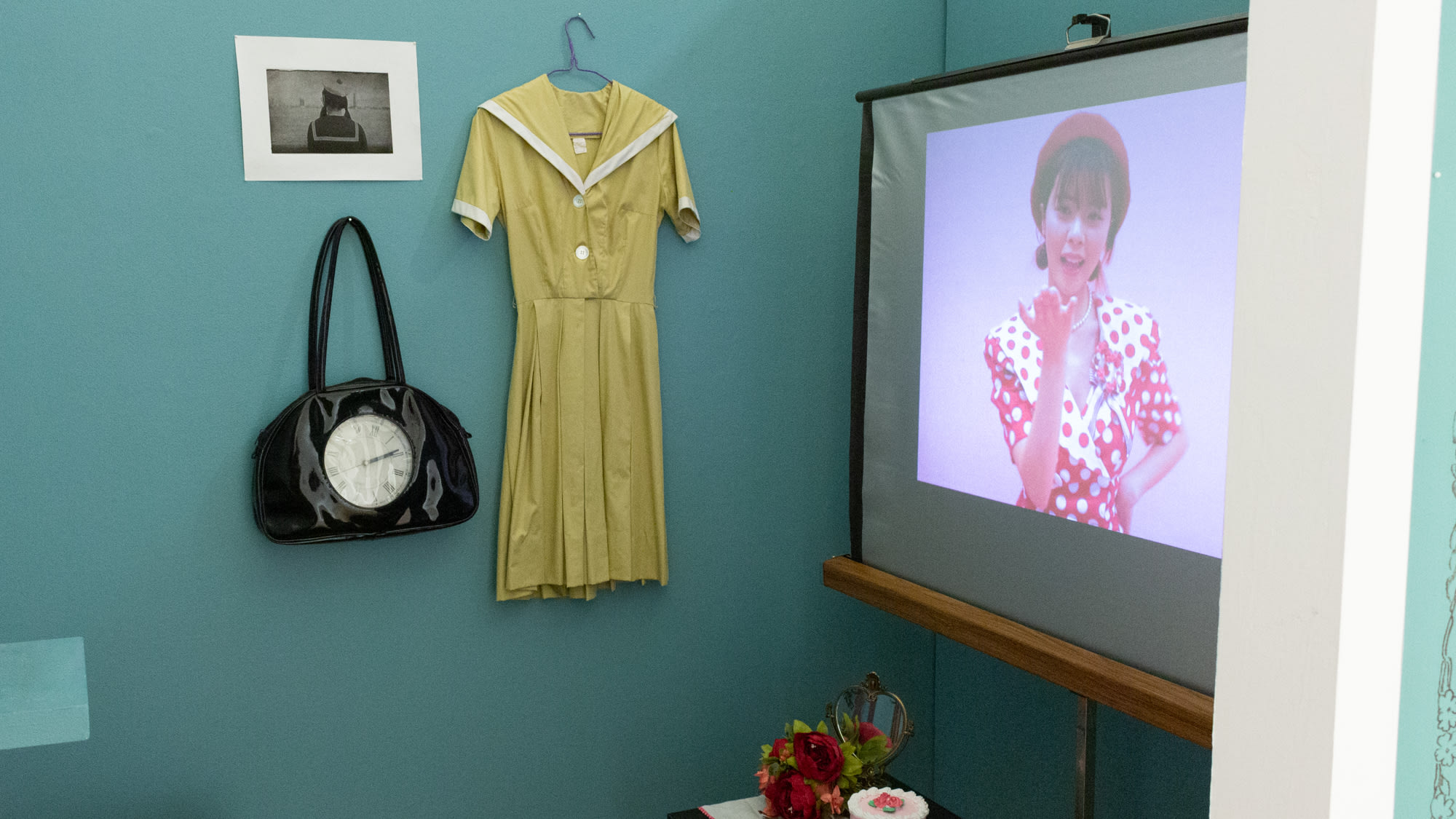 Photograph from inside partially enclosed installation space with green walls, a projector displaying a video of a sharply dressed woman, a yellow dress hanging on the wall, and other ephemera spread throughout space.