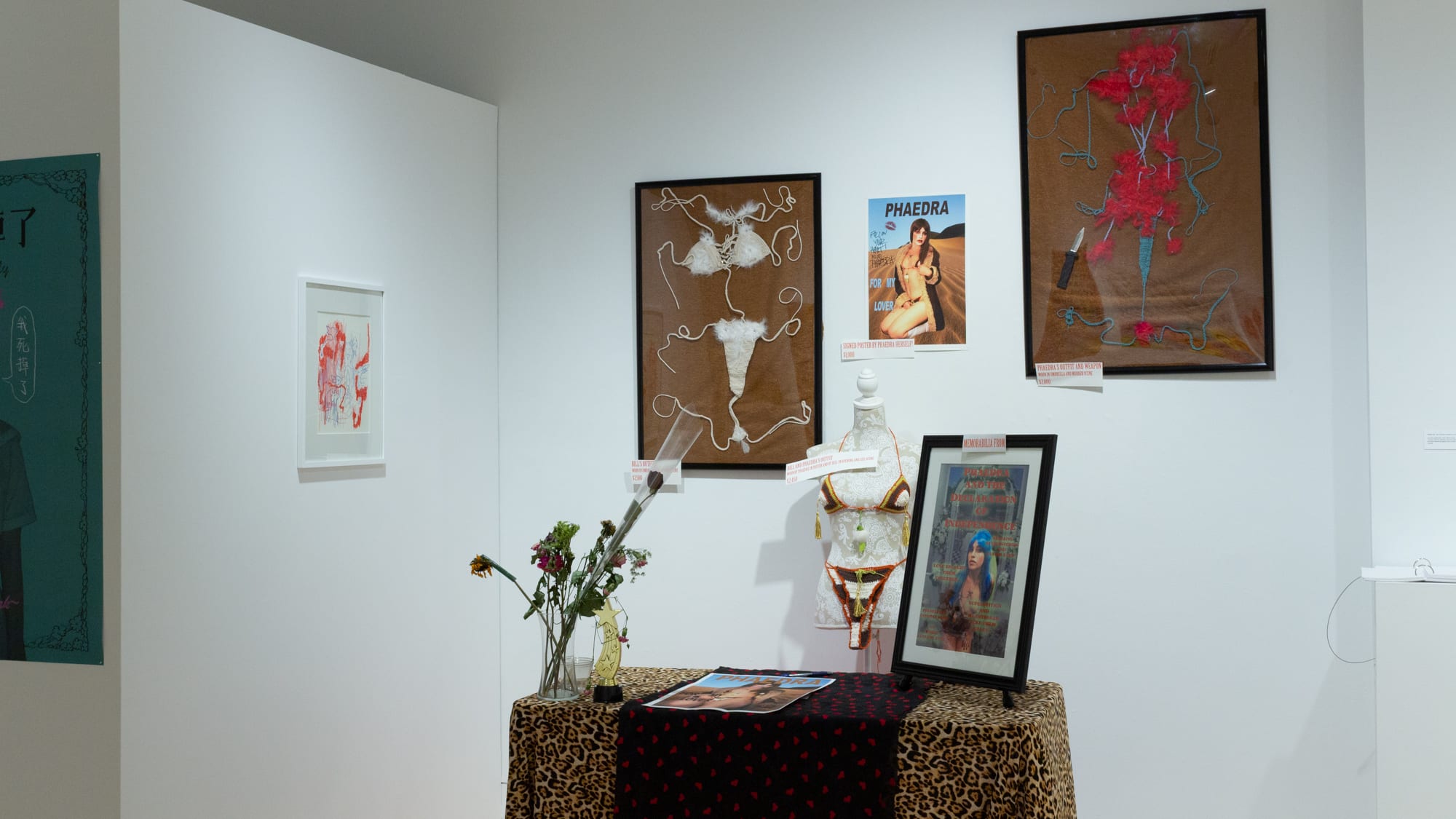 Installation containing multiple framed artworks of textile and promotional materials for an adult entertainer persona. In front of the framed works is a "booth"-like table with tablecloth and objects related to autograph signing.
