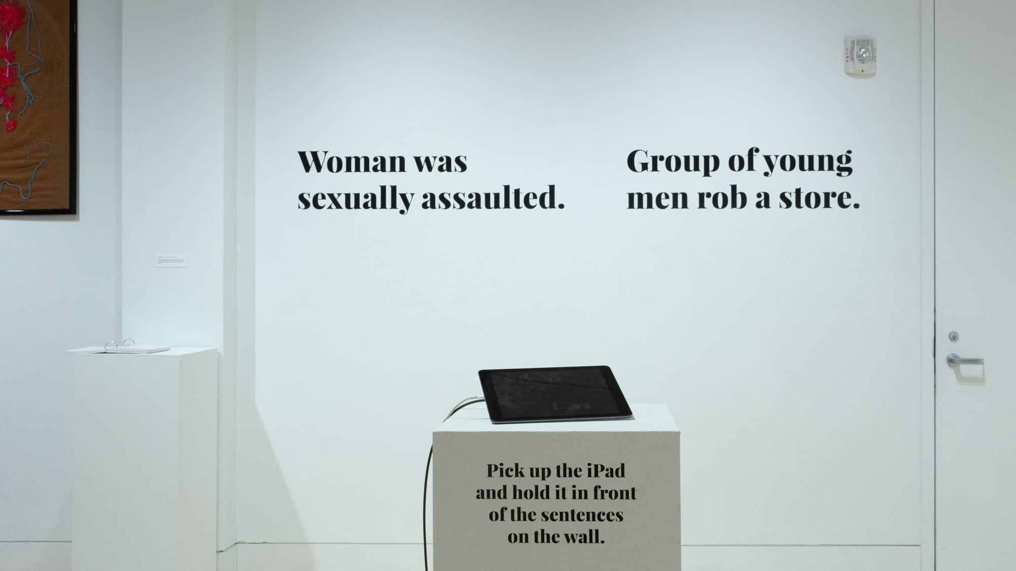 Photograph of wall in exhibition space, with text "woman was sexually assaulted" and "group of young men rob a store" written using black vinyl lettering. In front of them is an iPad on pedestal for a virtual reality experience.