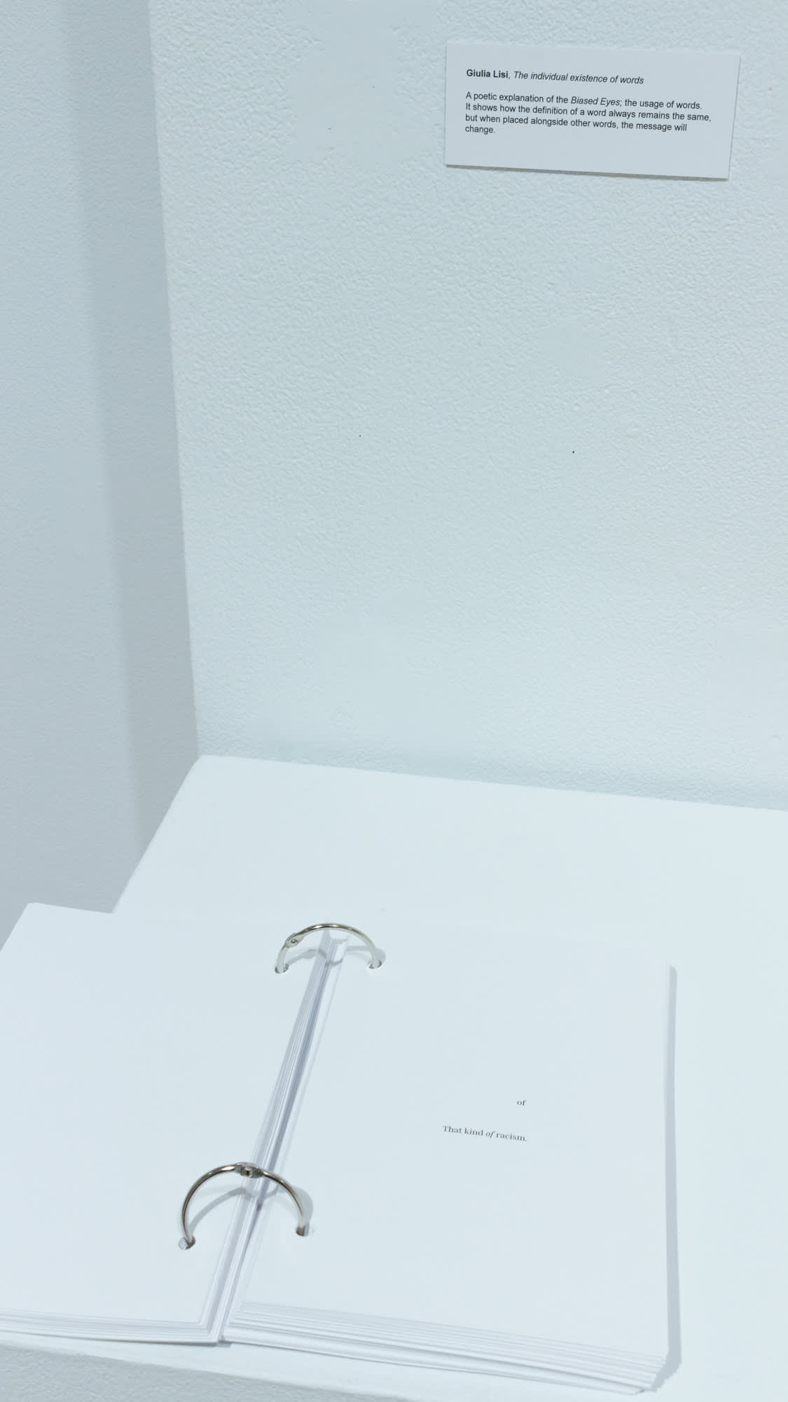 Close-up image of a book with text with the following text: "of" and "that kind of racism." Book is on pedestal with small label noting artist (Giulia Lisi) and small statement.