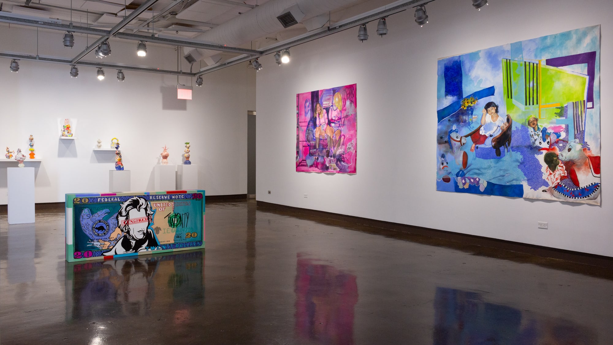 On the right is a large blue painting with a young girl looking at viewer, another painting by same artist in pink and purple is to its left, and on far left are multiple shelves and pedestals containing ceramic works. In the center of the space is a large wooden sculpture in a street art style depicting a $20 bill.