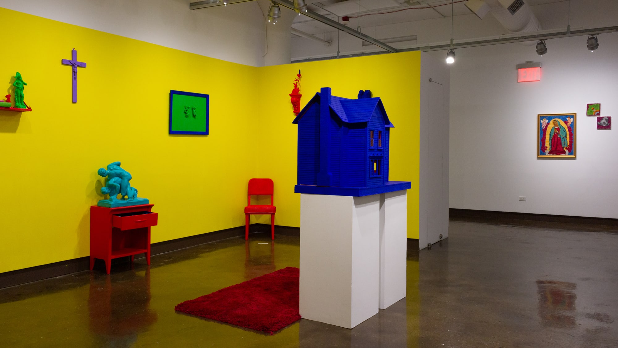 On the left is an inset space, painted yellow, with hyper-pigmented painted objects common to a household including a nightstand, chair, and large dollhouse. On the right are three small, colorful embroidery pieces on a nearby wall.